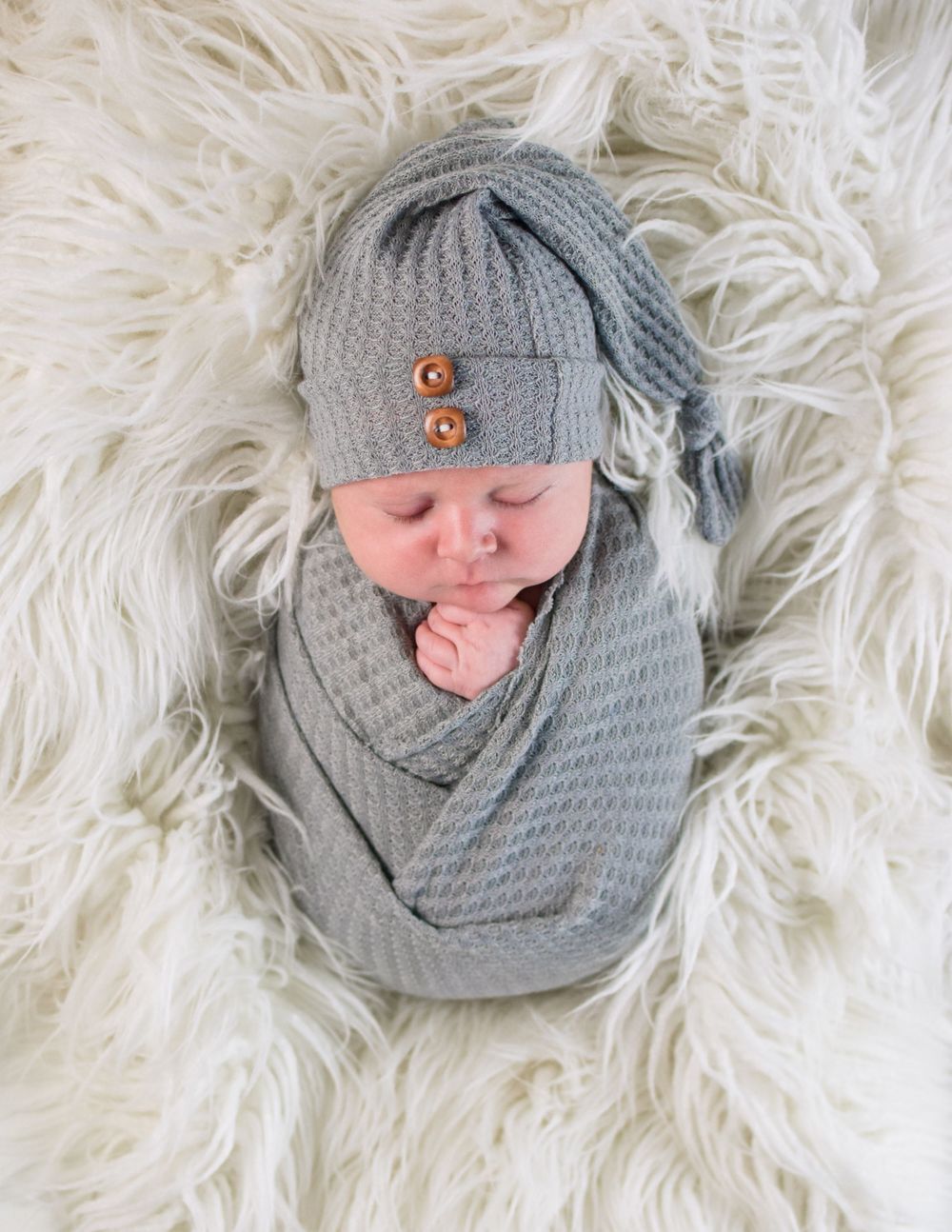 Newborn baby swaddled in gray knit wrap with matching knotted hat with wooden buttons on fluffy white fur backdrop.