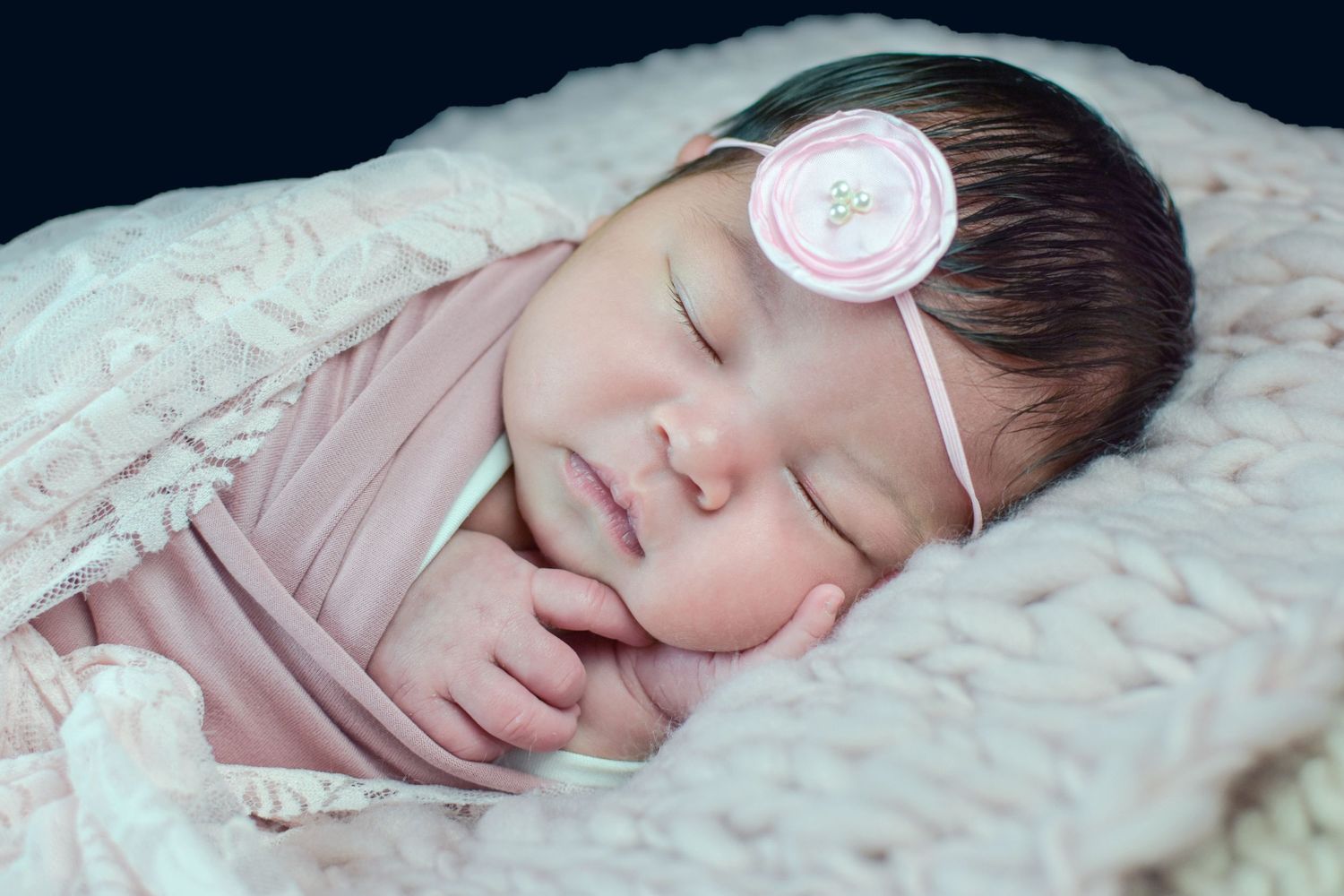 Peaceful newborn baby girl sleeping wrapped in pink, wearing a delicate floral headband on a soft blanket.