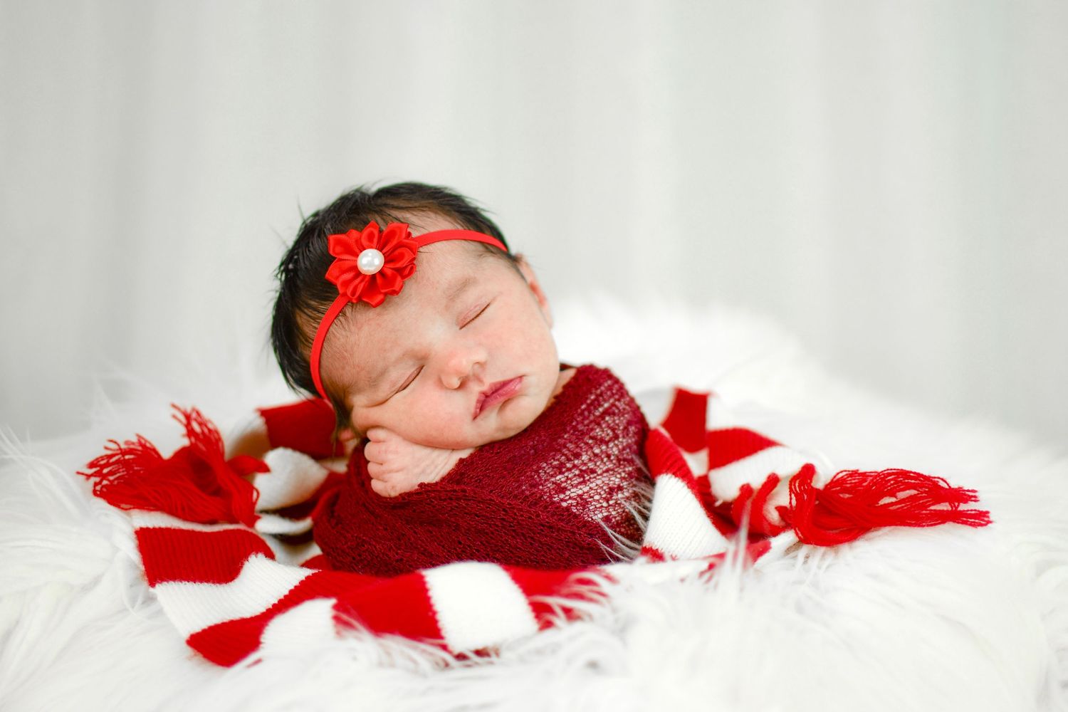 Sleeping newborn baby wrapped in red and white striped blanket with red flower headband on white fur.