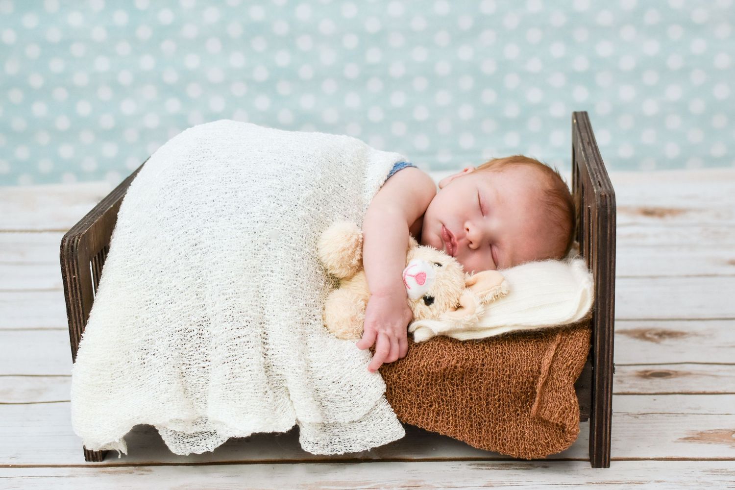 Newborn baby sleeping peacefully in a tiny wooden bed with a teddy bear and white blanket.