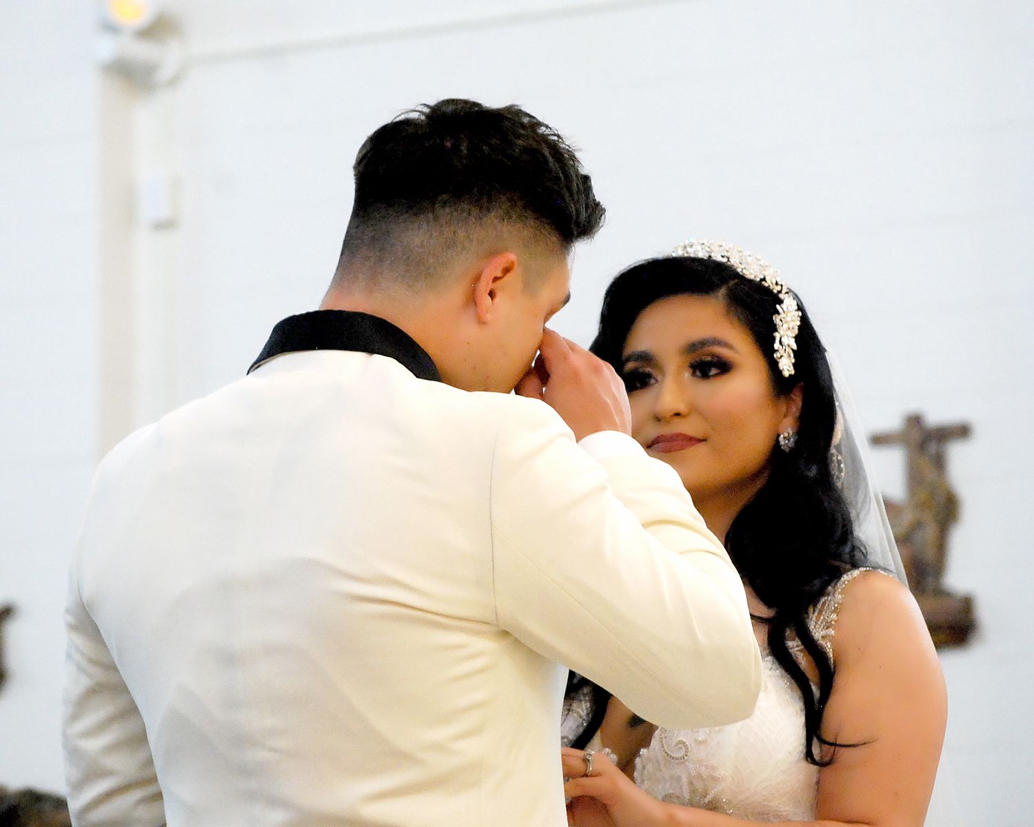 Groom wiping tears during emotional wedding ceremony while bride smiles tenderly in white gown and veil.