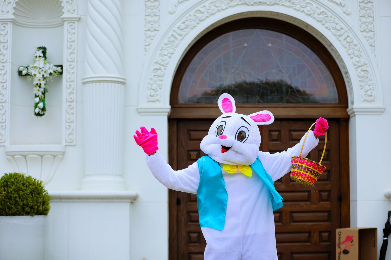 Easter bunny mascot in white costume with blue scarf holds basket outside decorated church with floral cross.