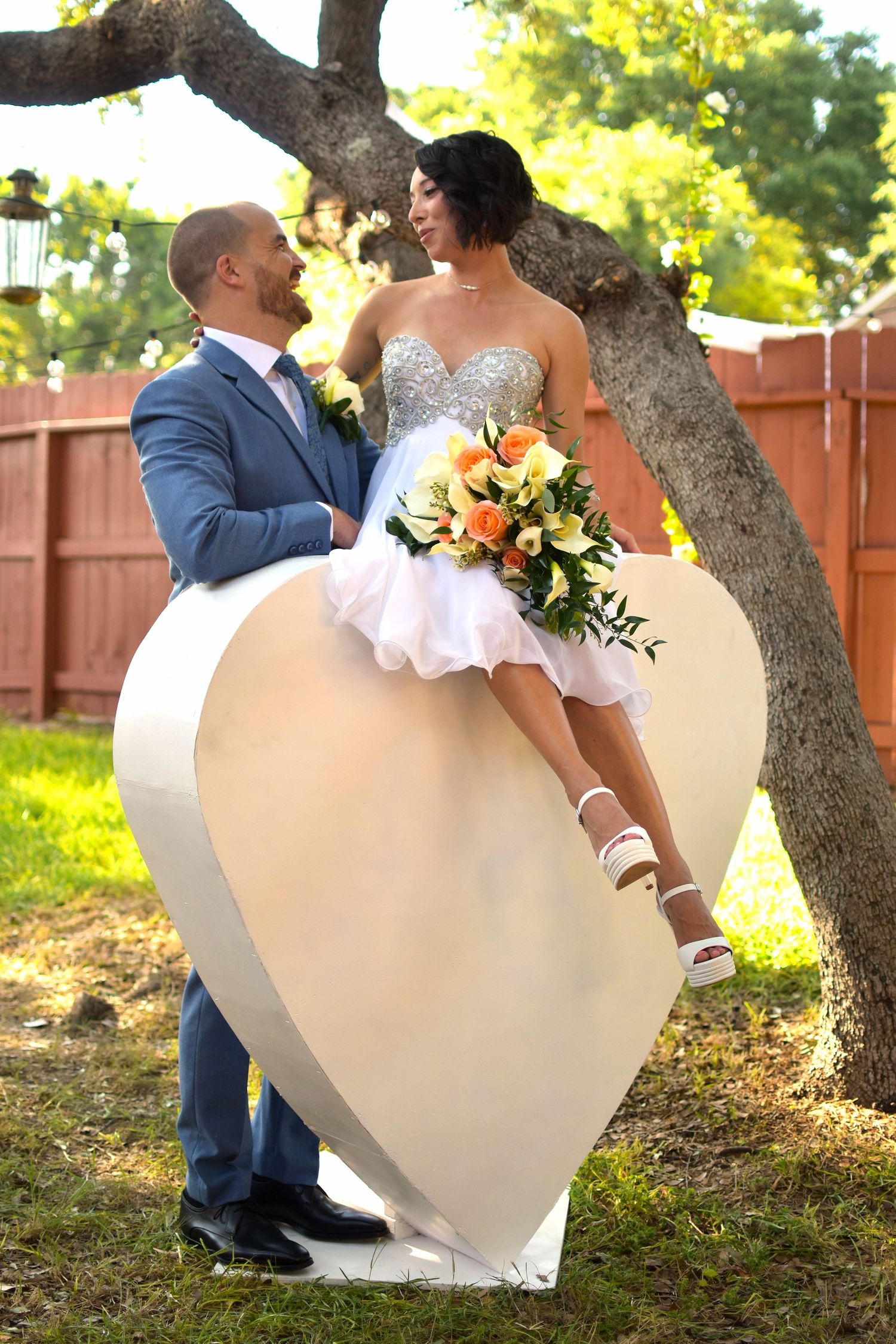 Bride sitting on large heart prop held by groom in blue suit outdoors, holding orange and yellow flower bouquet.