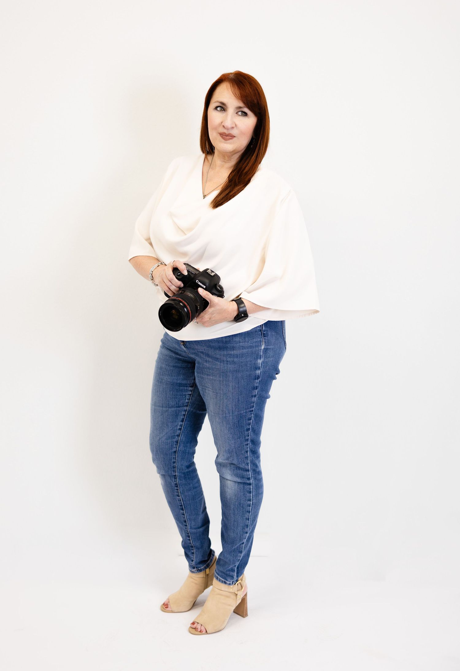 A woman with red hair holds a DSLR camera, wearing a white top and blue jeans, posing on a white background.