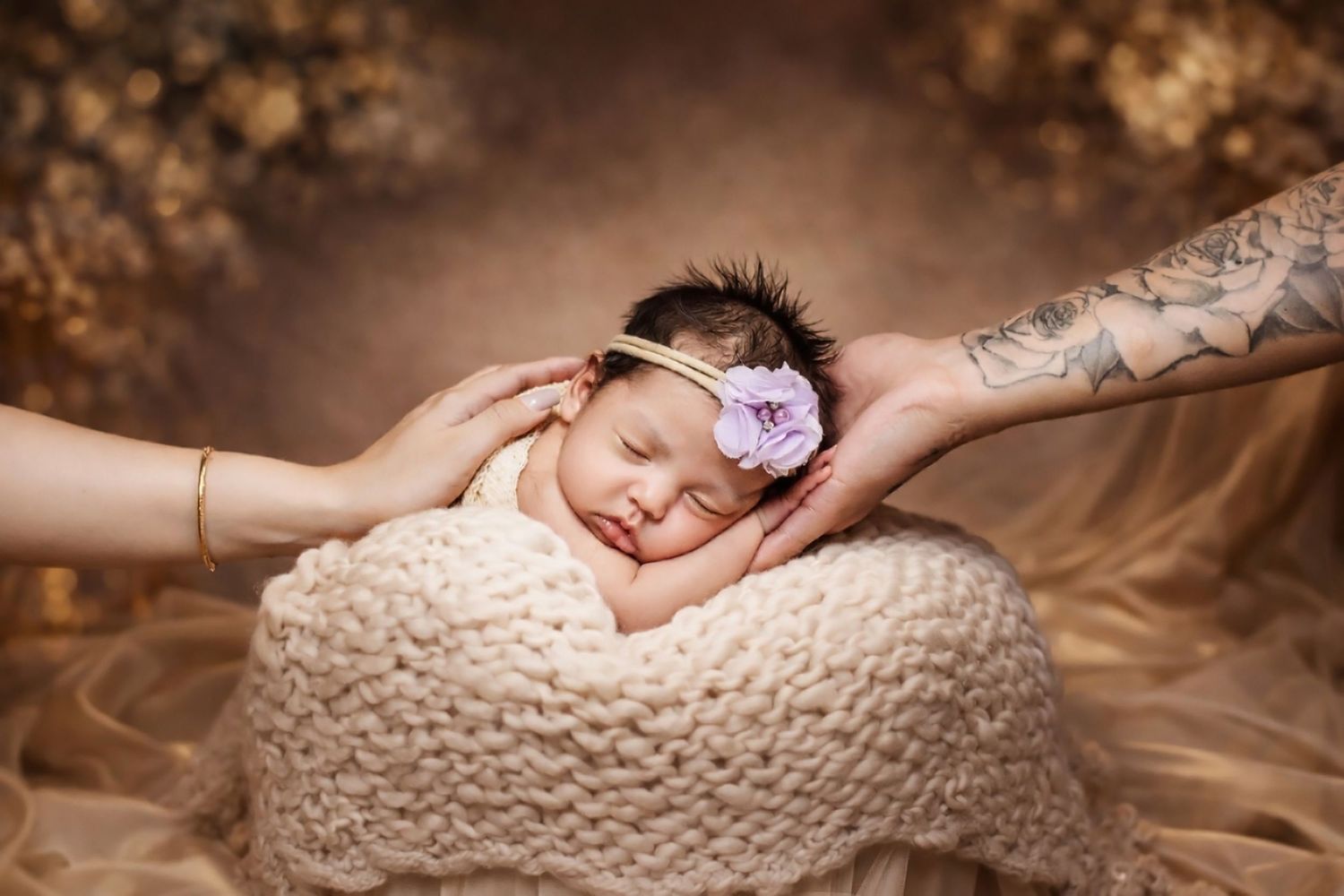 Newborn baby girl sleeping peacefully in a knit bowl with a pink flower headband, parents' hands gently cradling her head.