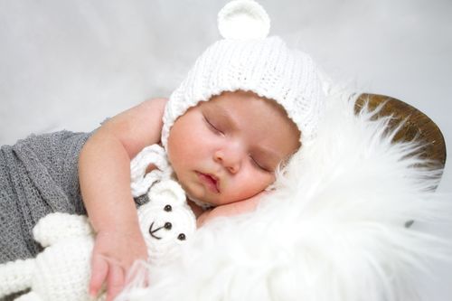 Sleeping newborn baby wearing a white knit bear hat, cuddling a small white teddy bear on a fluffy white blanket.