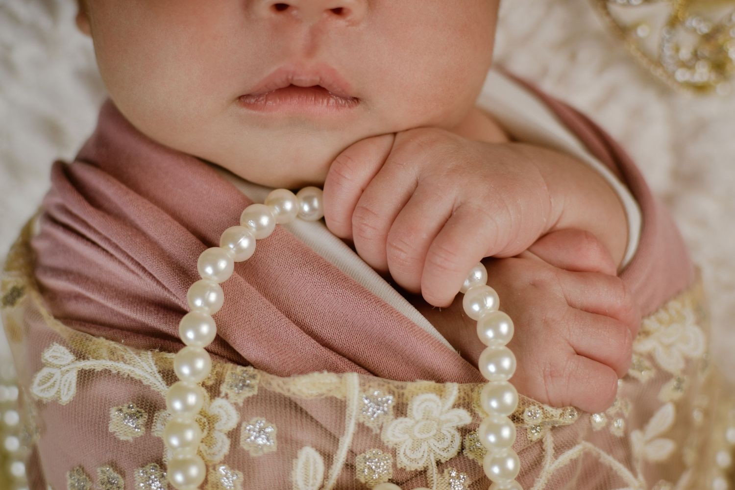 Close-up of a newborn baby in pink lace outfit holding a pearl bracelet with tiny delicate fingers.