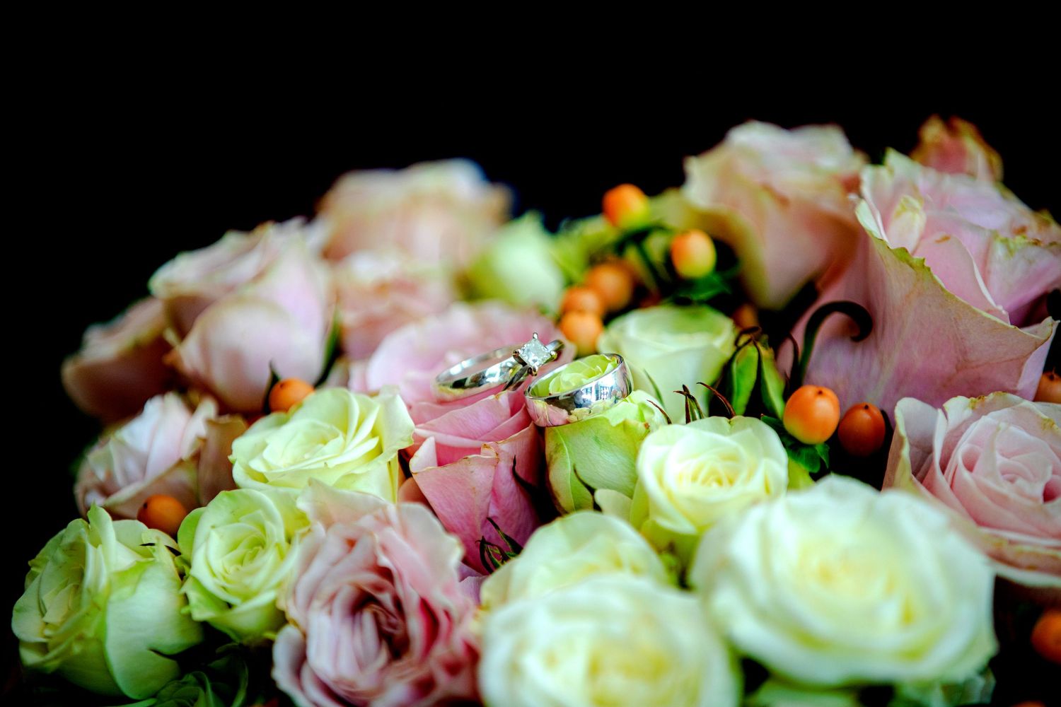 Two silver wedding rings rest on a lush bouquet of pink and white roses with orange berries.