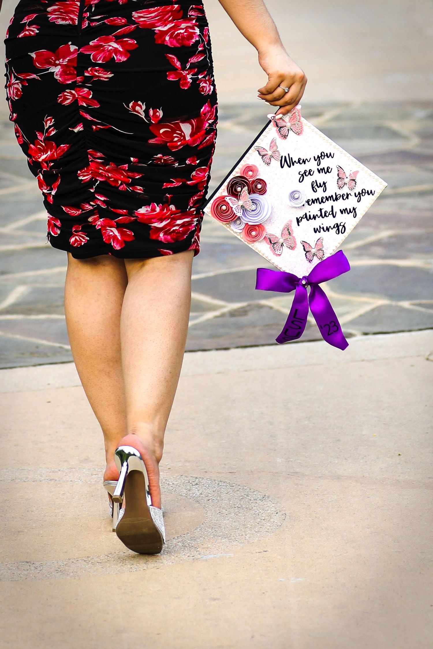 Woman in floral skirt walking while holding decorated graduation cap from LSU with purple bow at her graduation photo session Conception Pk