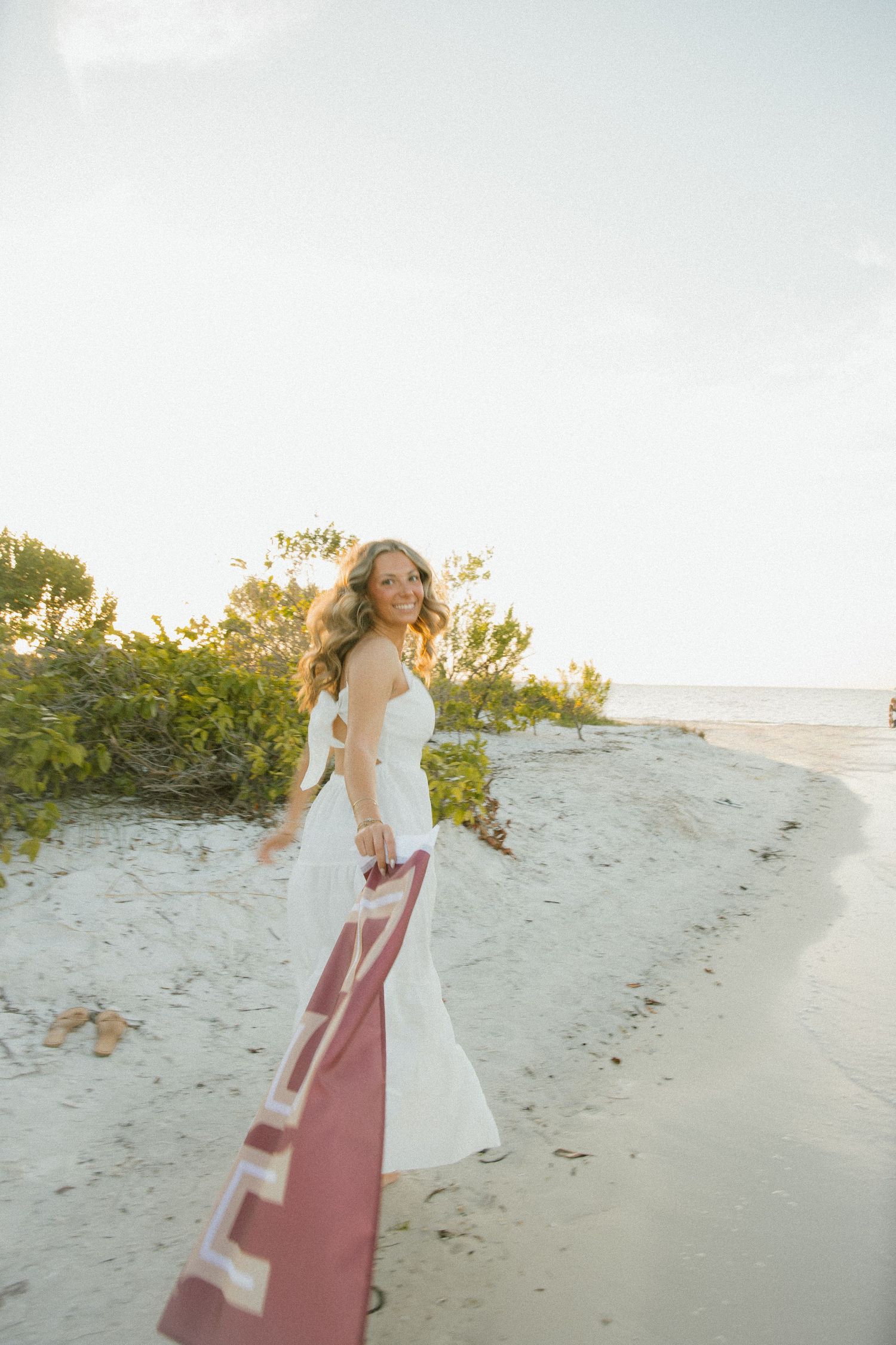 Woman in a white dress looks back and smiles while carrying an FSU flag along a sandy shoreline at sunset.