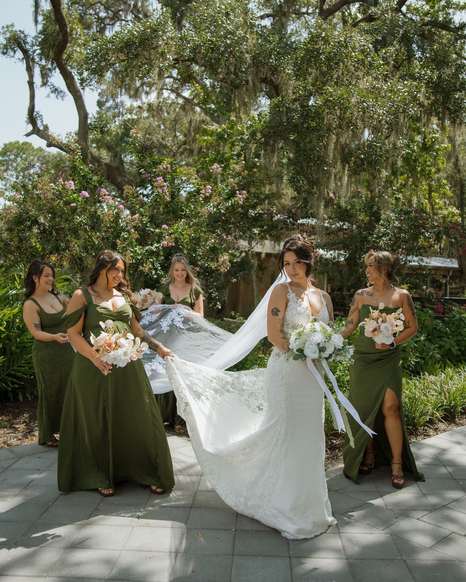 Bride in a lace wedding gown with a flowing veil poses with four bridesmaids in olive green dresses holding bouquets under mossy oak trees.
