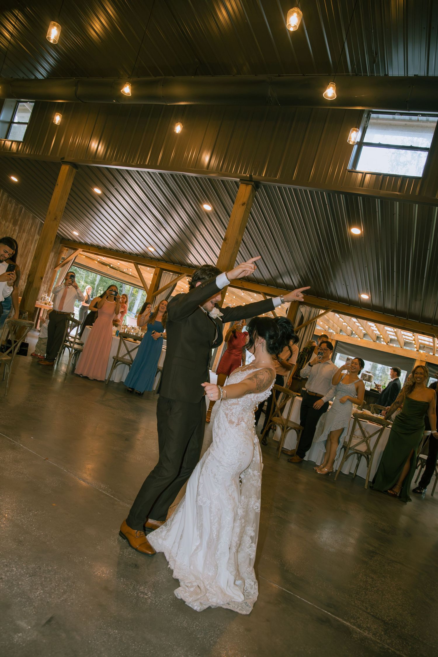 Bride in a lace gown dances with her groom in a rustic barn venue while wedding guests watch from the sides.