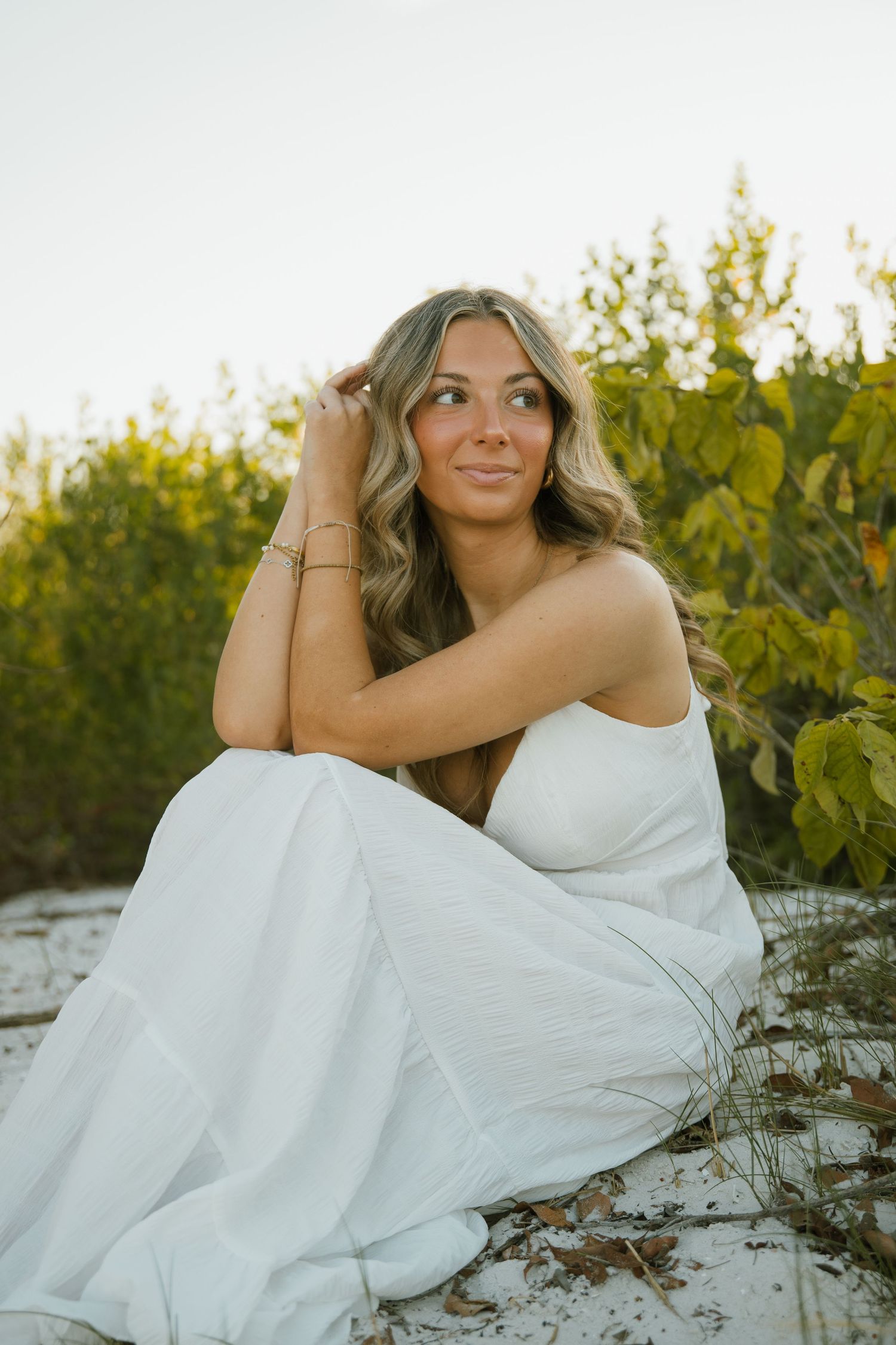 Woman with long wavy hair in a white dress sits on the sand with a thoughtful smile in front of green foliage.