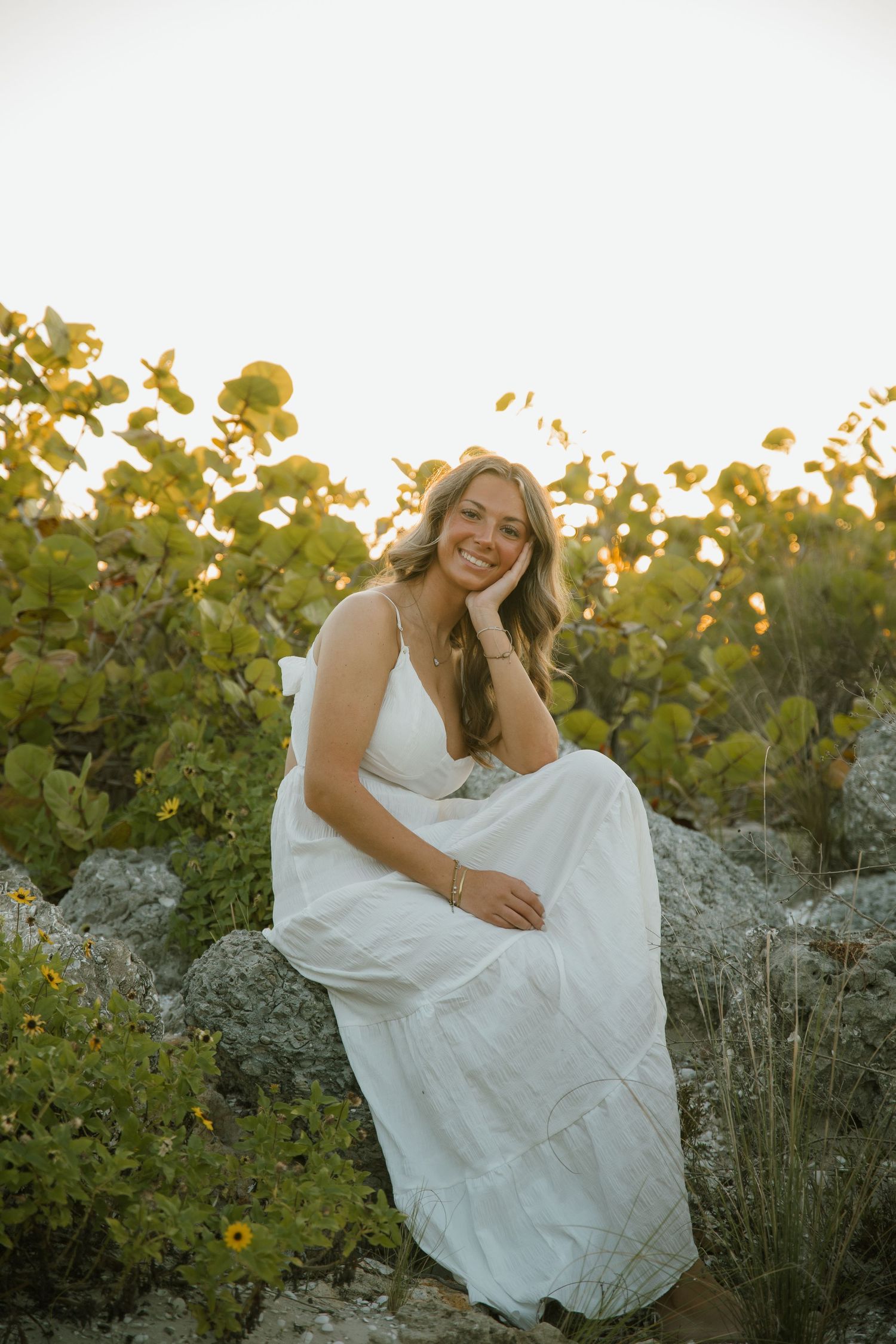 Smiling woman in a white sundress sits on coastal rocks surrounded by green sea grape leaves at sunset.