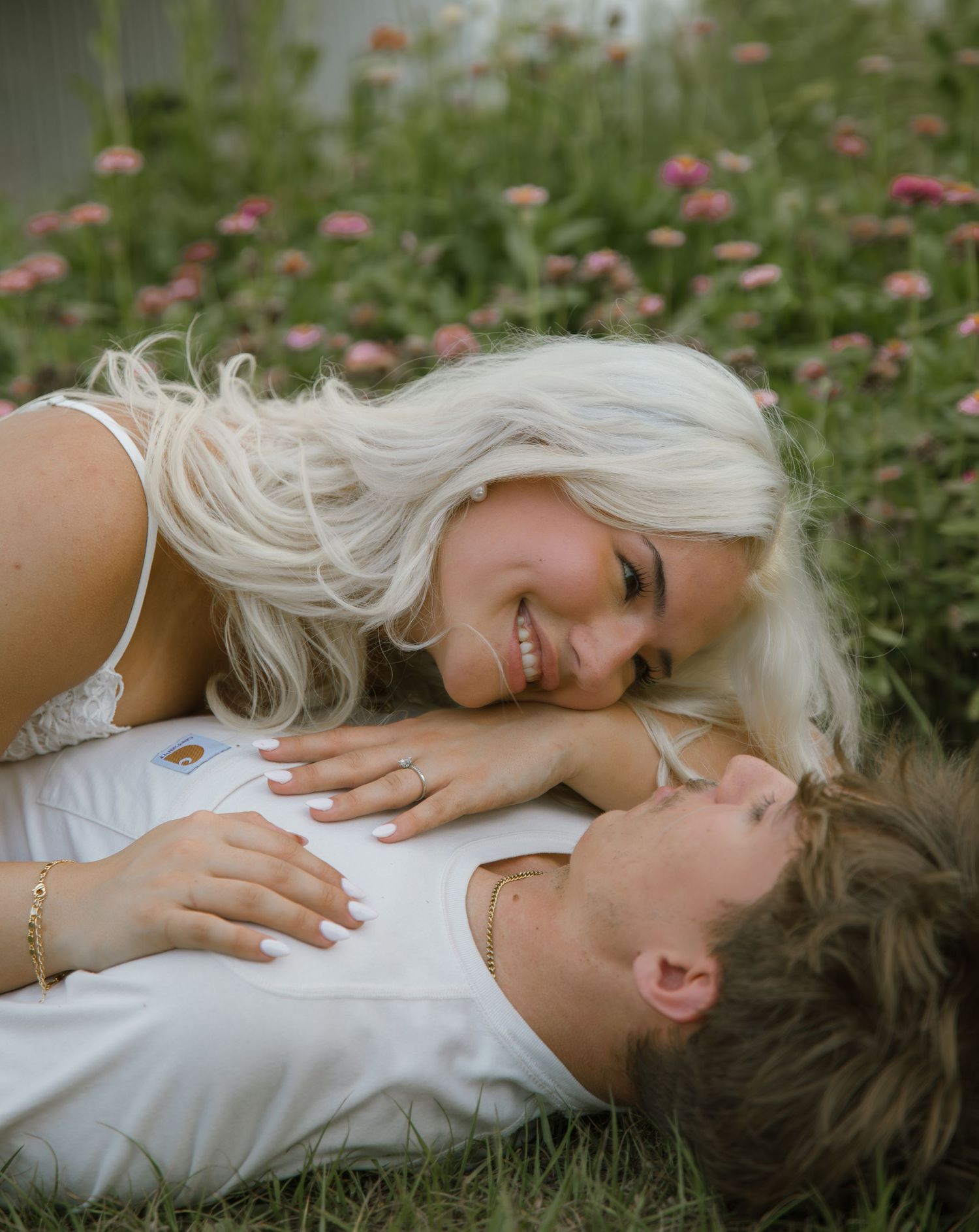 Smiling blonde woman leaning over a man lying in the grass near pink wildflowers in a romantic couple photo.