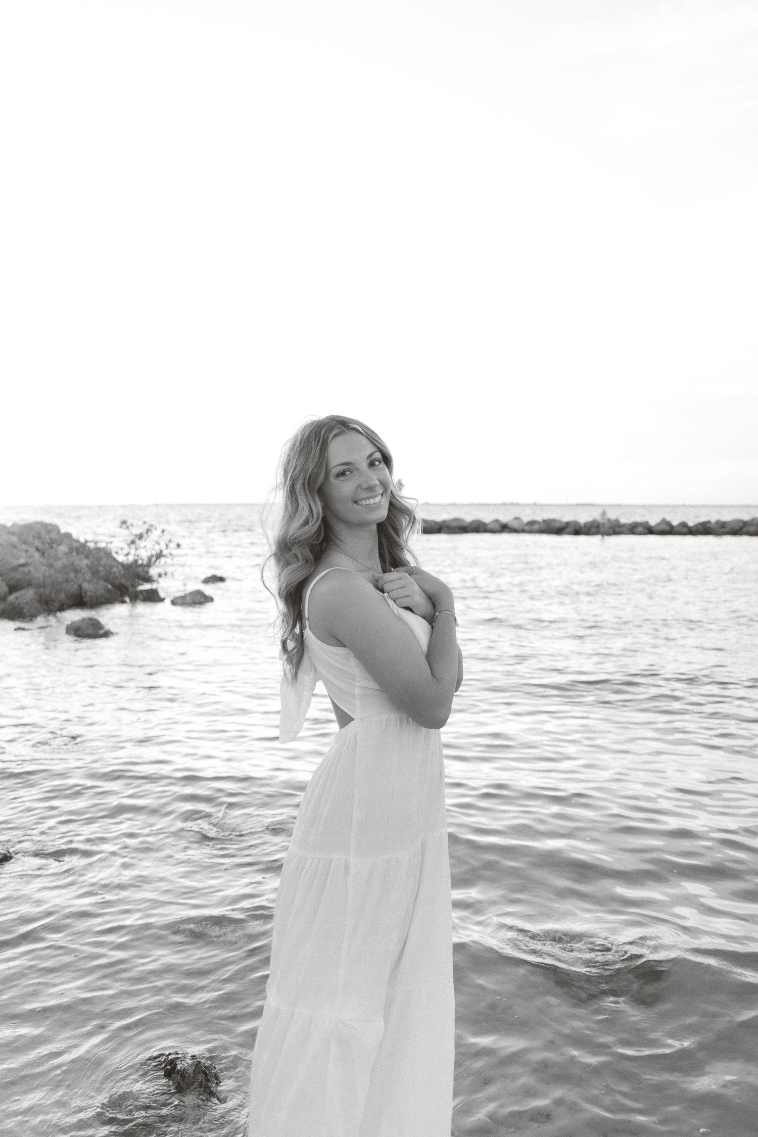 Black and white portrait of a smiling woman in a long white dress standing in shallow ocean water.