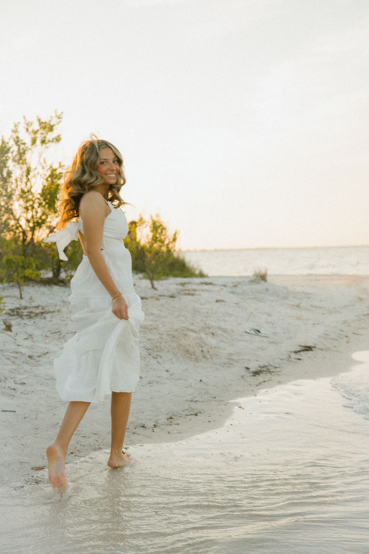 Smiling woman in a white sundress walks barefoot along a sandy shoreline at sunset, glancing back over her shoulder.