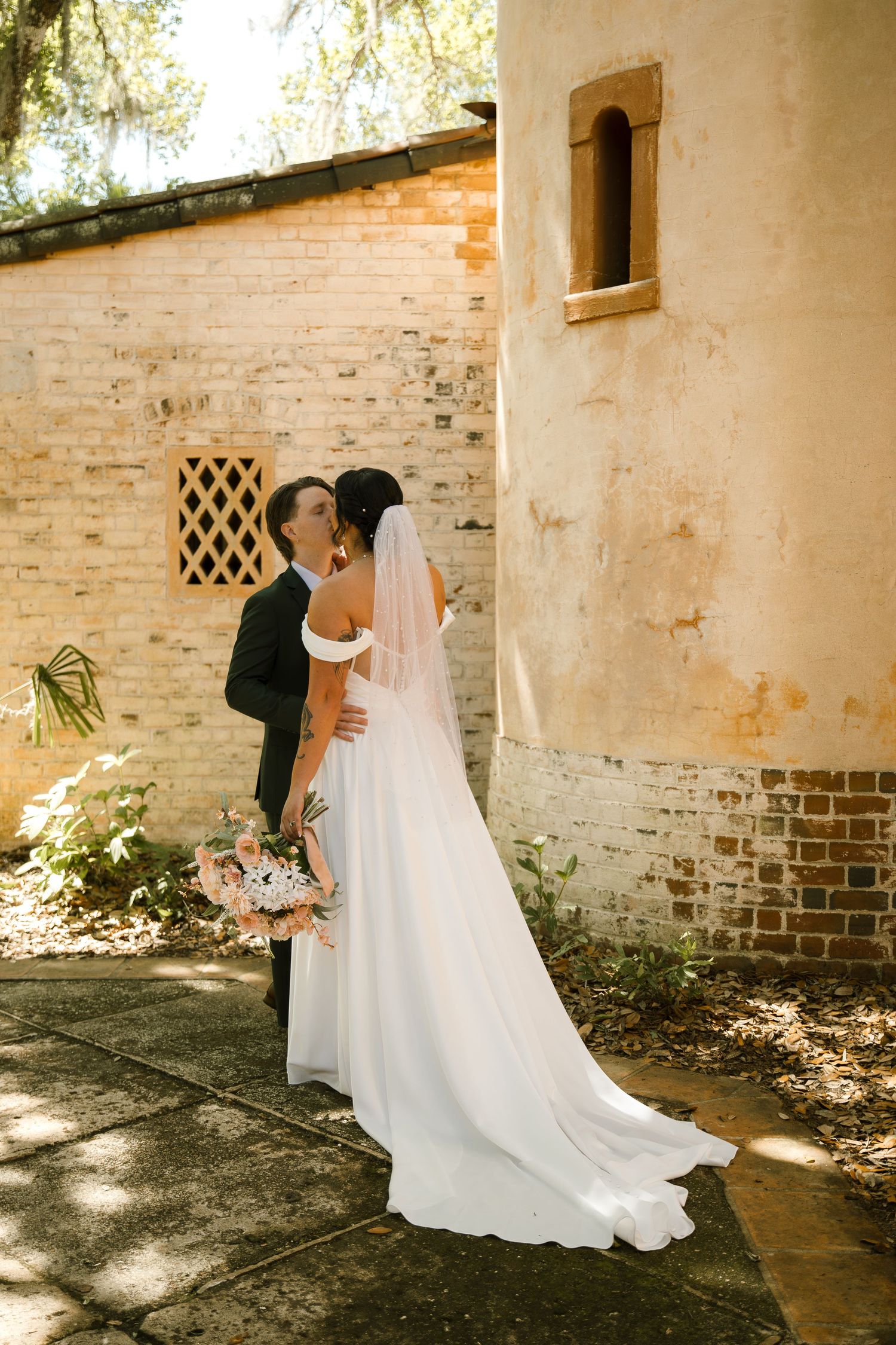 Bride in a flowing white gown and veil kisses her groom beside a historic brick building holding a bouquet.