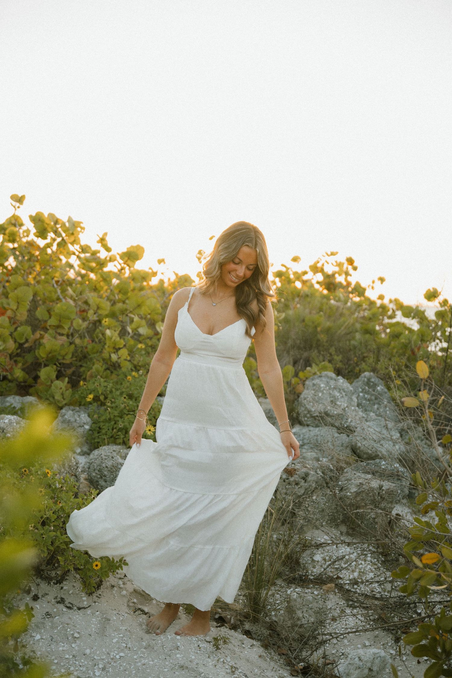 Barefoot woman in a flowing white maxi dress holds her skirt and smiles down while standing on rocks near sunlit coastal shrubs.
