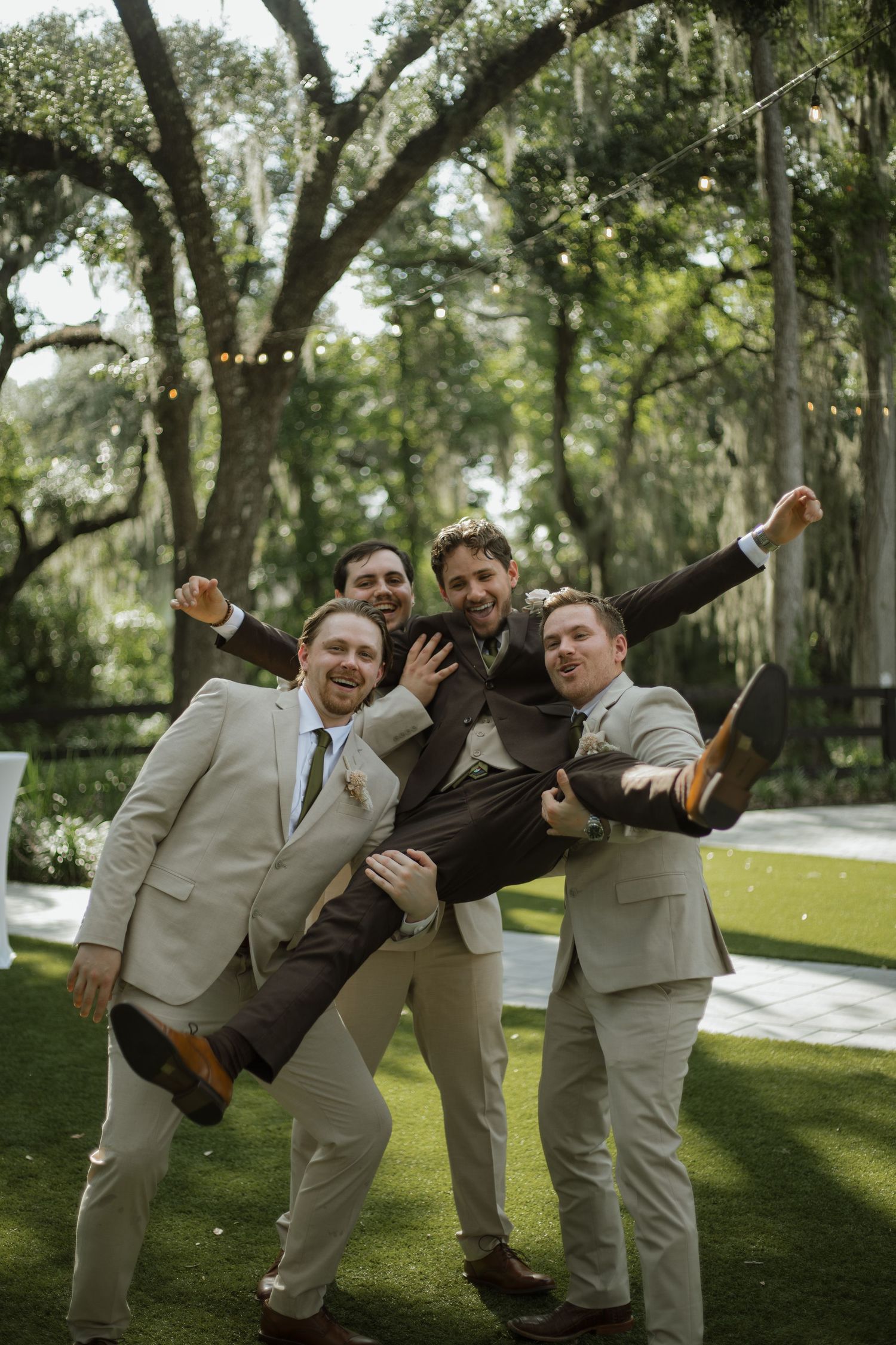 Groomsmen in tan suits playfully lift the groom into the air on a lawn beneath oak trees with string lights.