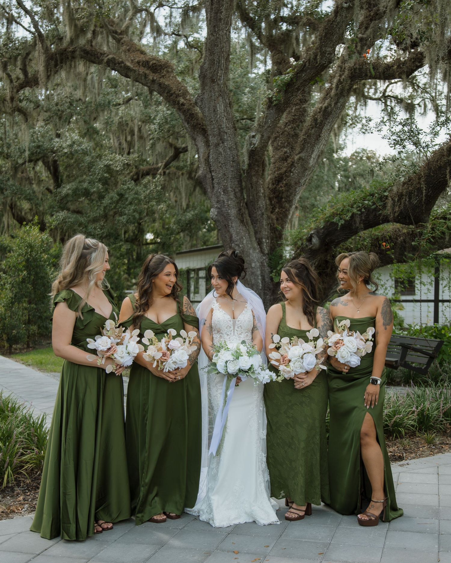 Bride in a white gown stands among four bridesmaids in olive green dresses holding white bouquets beneath a moss-draped oak tree.