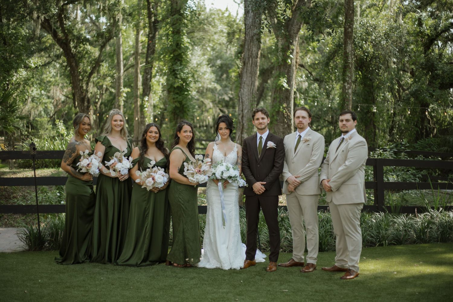 Bride in white gown with four bridesmaids in green dresses and three groomsmen in suits pose together on a lawn at an outdoor wedding.