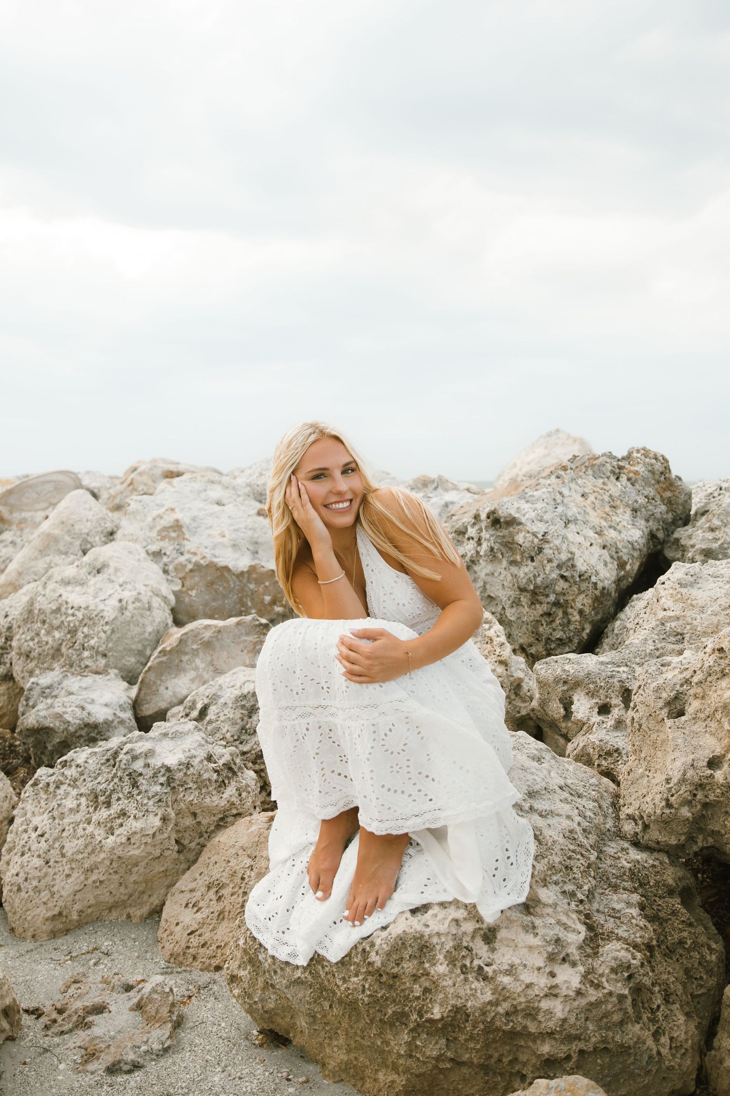 Smiling woman with long blonde hair in a white eyelet dress sits barefoot on large coastal rocks under a soft, cloudy sky.