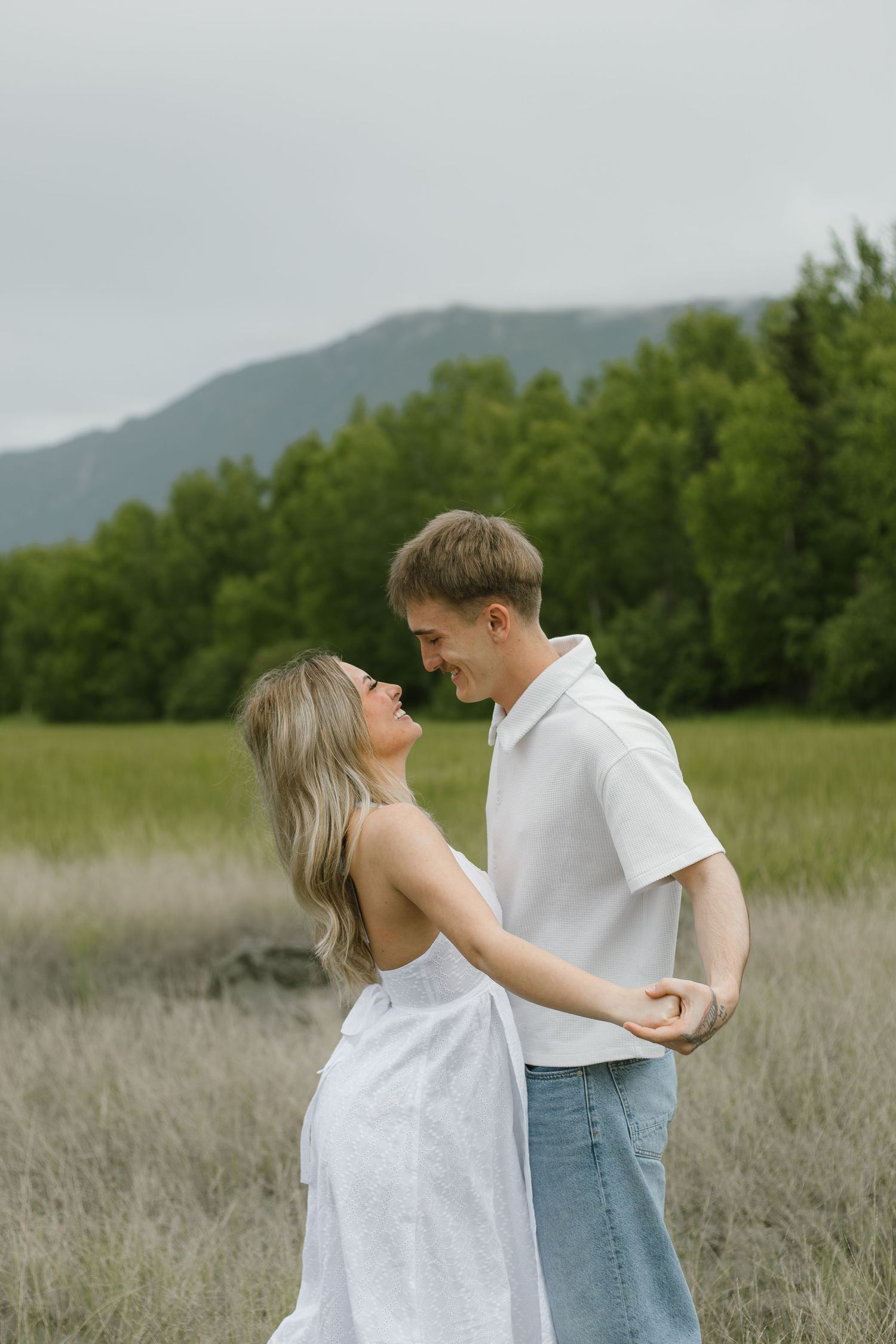 Smiling couple in white outfits dance and hold hands in a grassy meadow with green trees and mountains in the background.