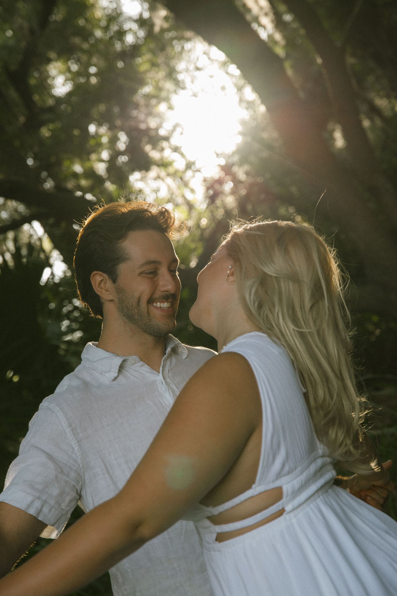 Smiling couple embraces face to face in dappled golden sunlight under trees during an outdoor engagement photo session.