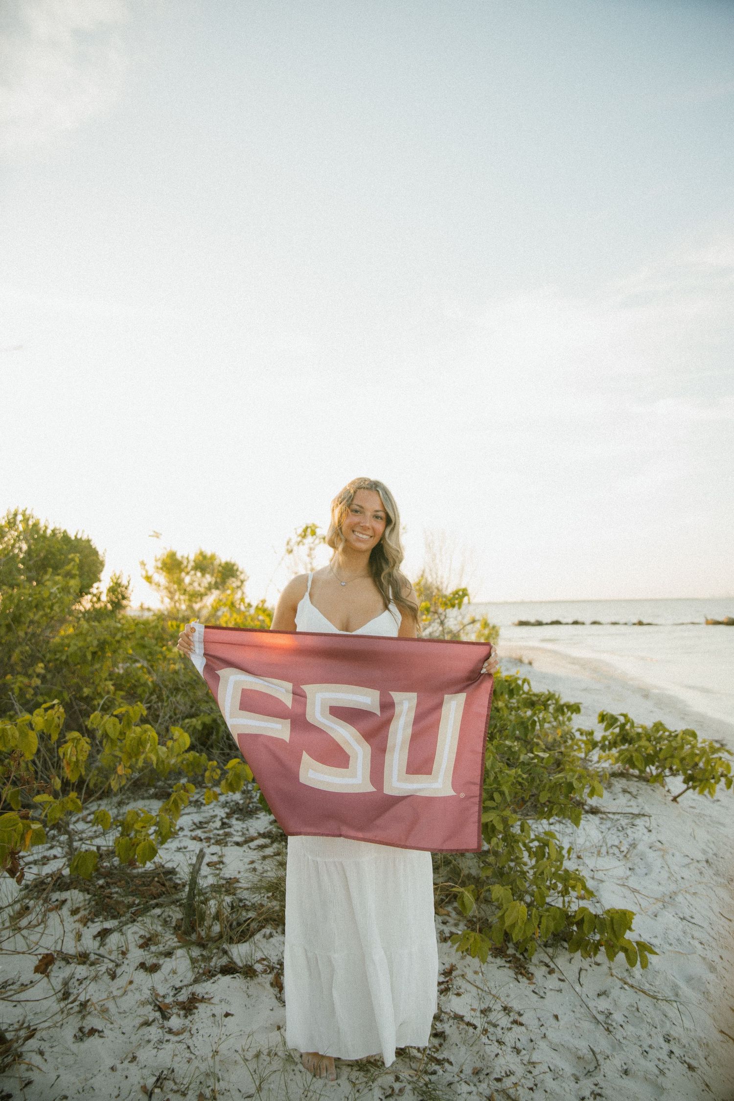 Smiling woman in a white dress holds a garnet FSU flag on a sandy beach surrounded by coastal greenery.