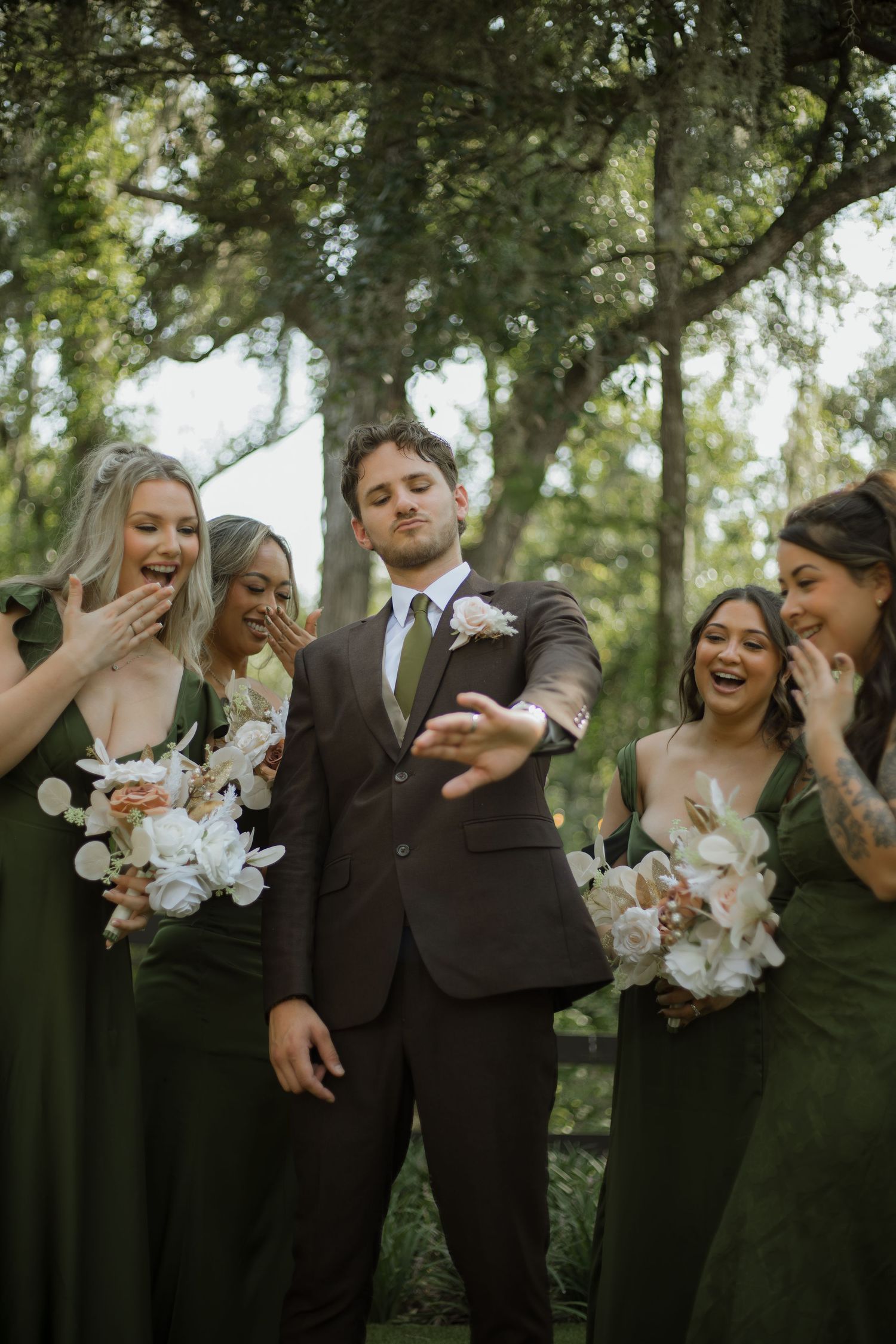 Groom in a brown suit shows his hand as four bridesmaids in green dresses with white floral bouquets laugh and react outdoors.