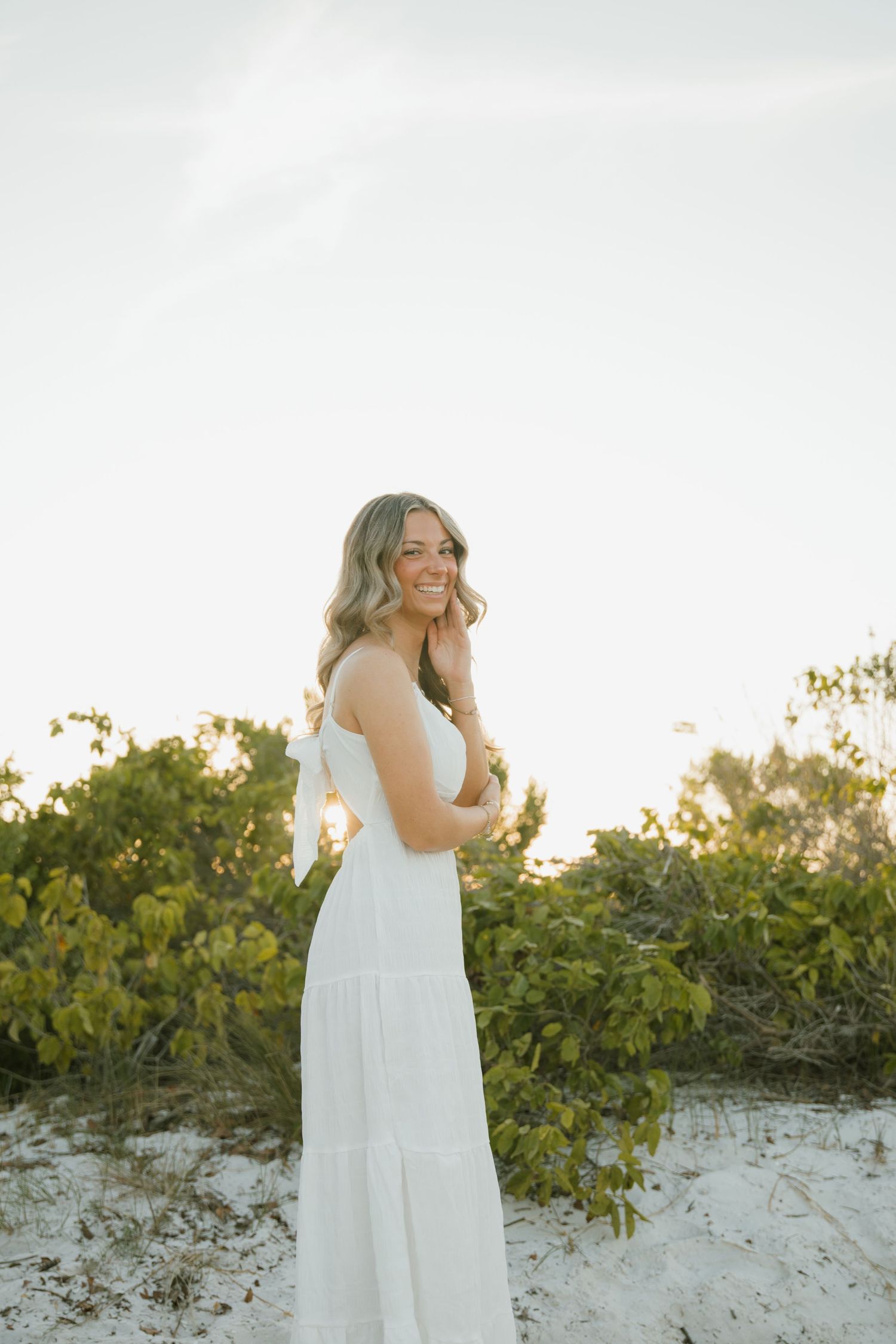 Woman in a flowing white dress smiles with her hand near her face on a sandy beach dune at sunset.