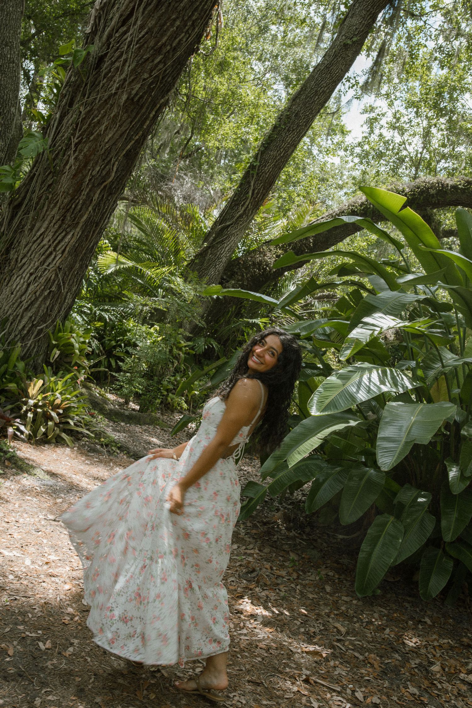 Woman in a flowing white floral maxi dress twirls and smiles on a shaded dirt path surrounded by tall trees and lush tropical plants.