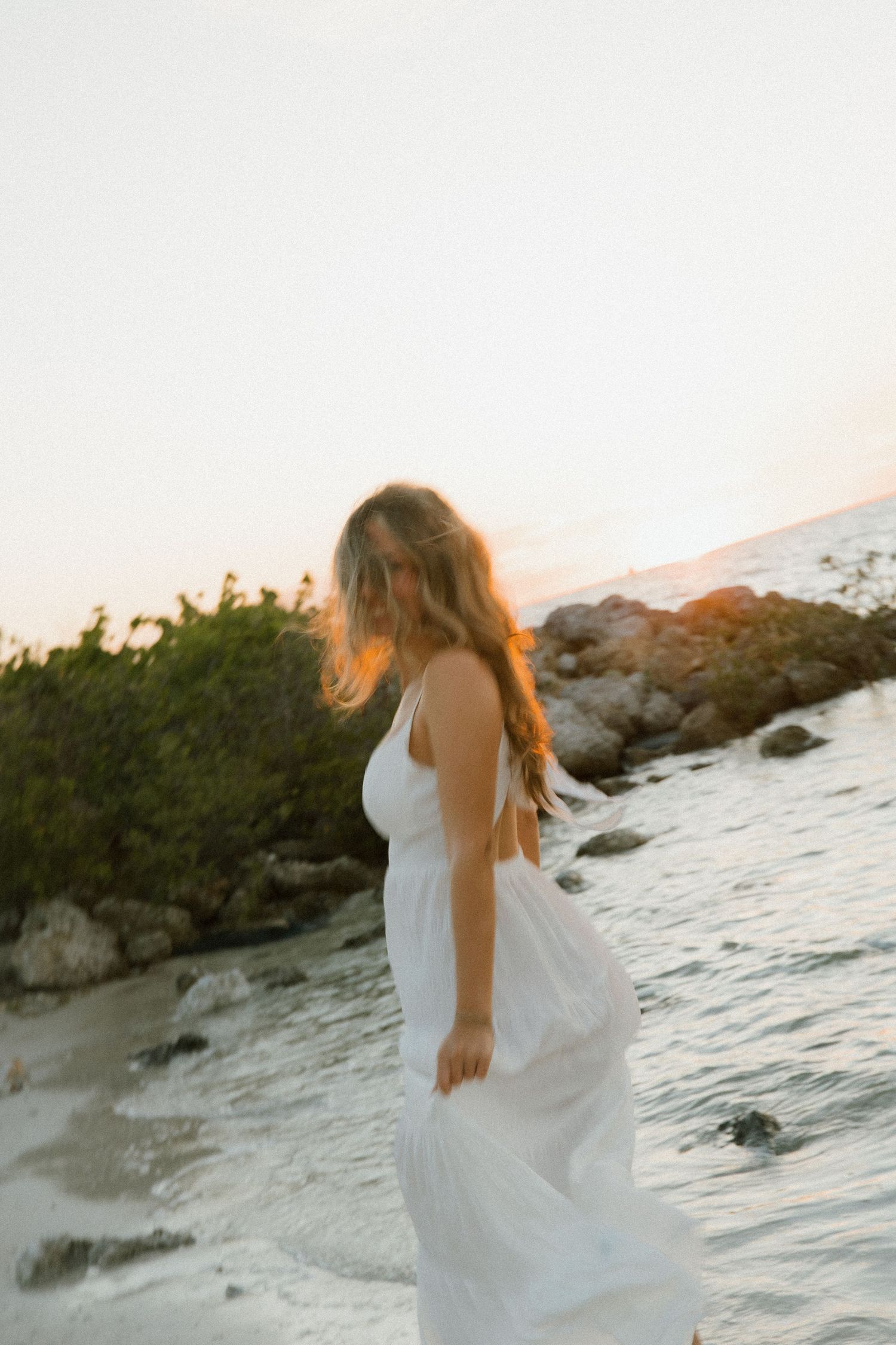 Woman in a white dress with wavy hair stands at the rocky water's edge as golden sunset light glows behind her.