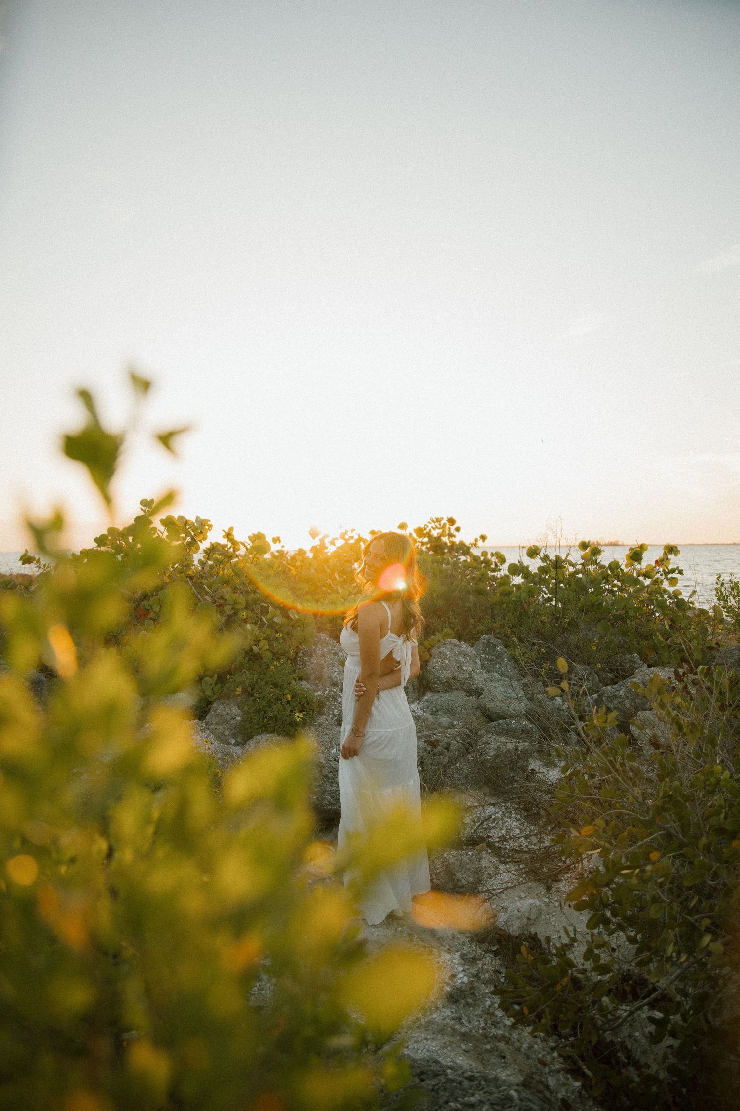 Woman in a white dress stands among seaside rocks and glowing green foliage as the golden sunset lights the horizon.