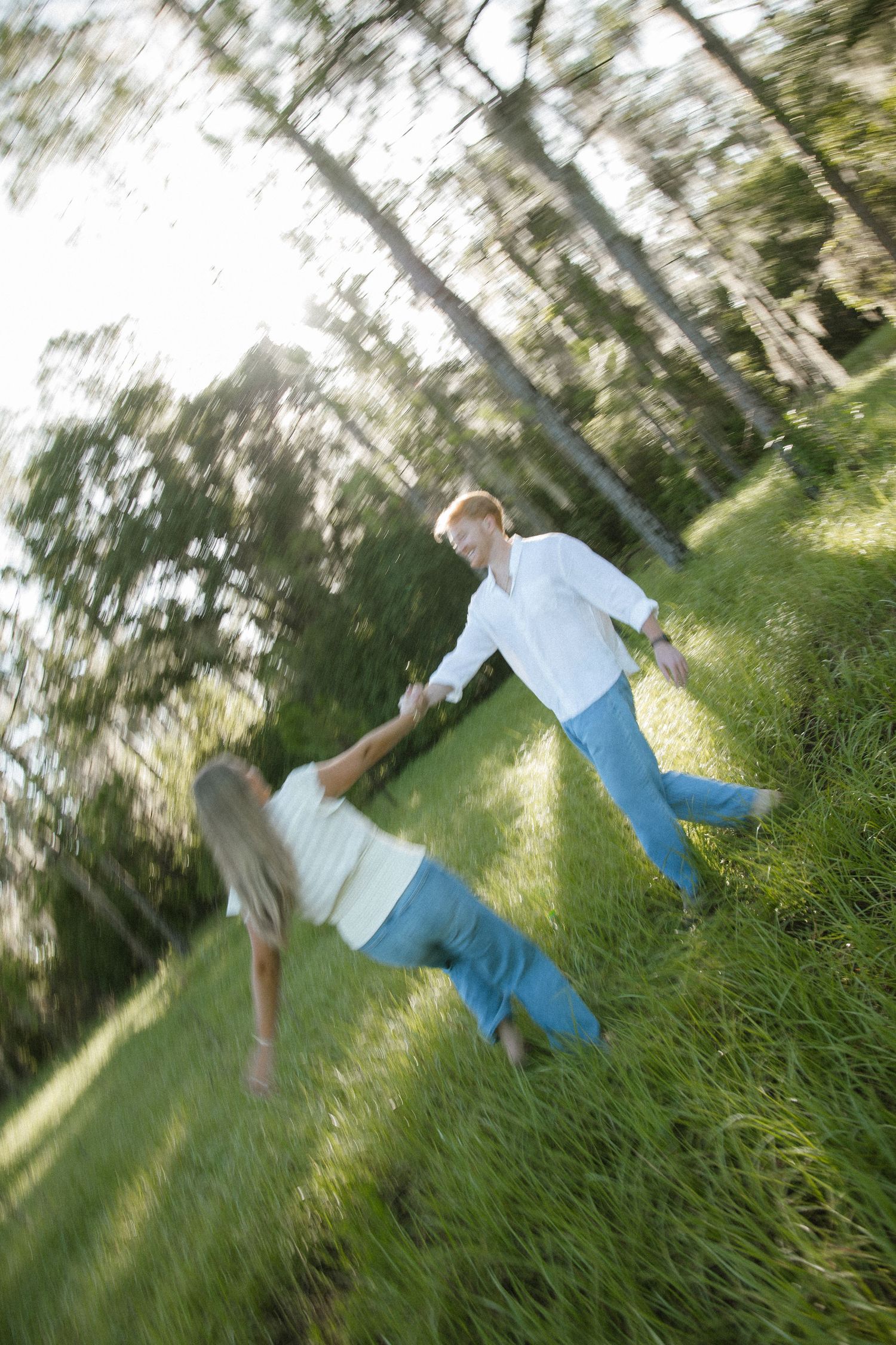 A man and woman hold hands and spin playfully on a sunlit grassy hillside surrounded by tall trees.
