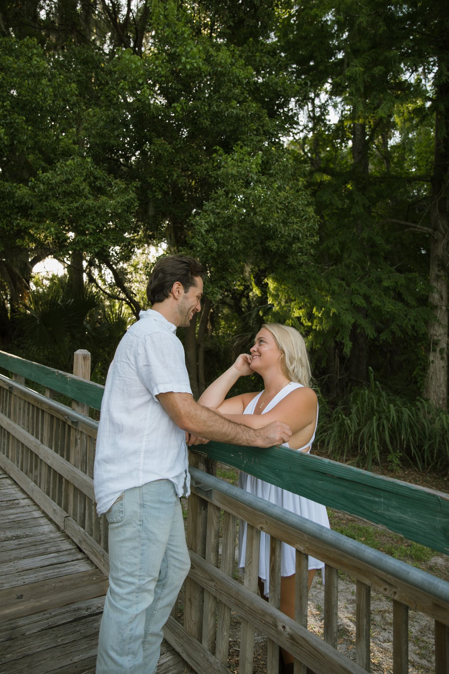 Smiling couple share a tender moment as they lean on a green wooden bridge railing surrounded by lush trees.