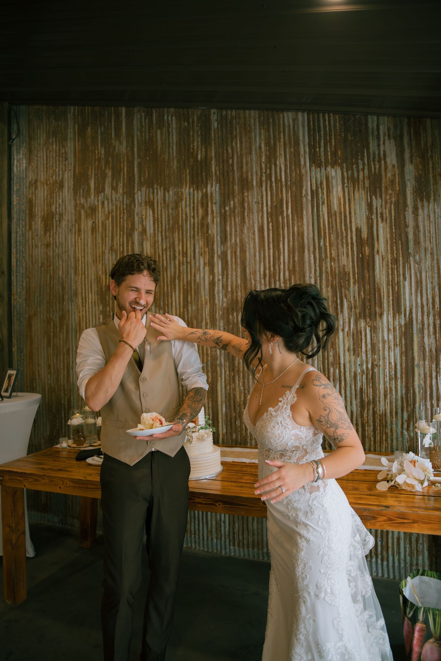 Bride in a lace dress playfully smears cake on the groom's face as he laughs holding a plate of wedding cake.
