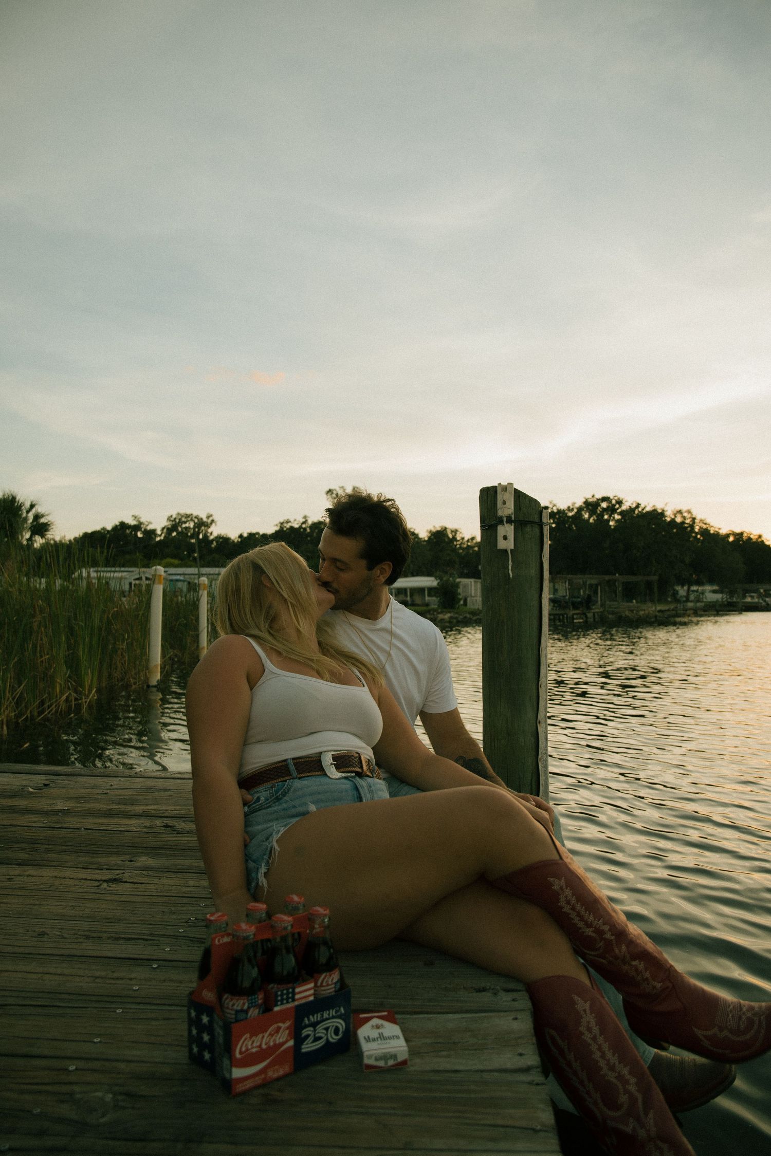 Couple kissing on a wooden lakeside dock at dusk with a six-pack of Coca-Cola bottles and cowboy boots.