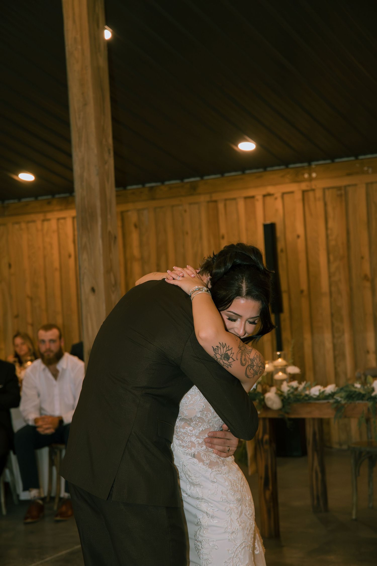 Bride in a lace gown and groom in a dark suit share an emotional embrace inside a rustic wooden barn venue with guests nearby.