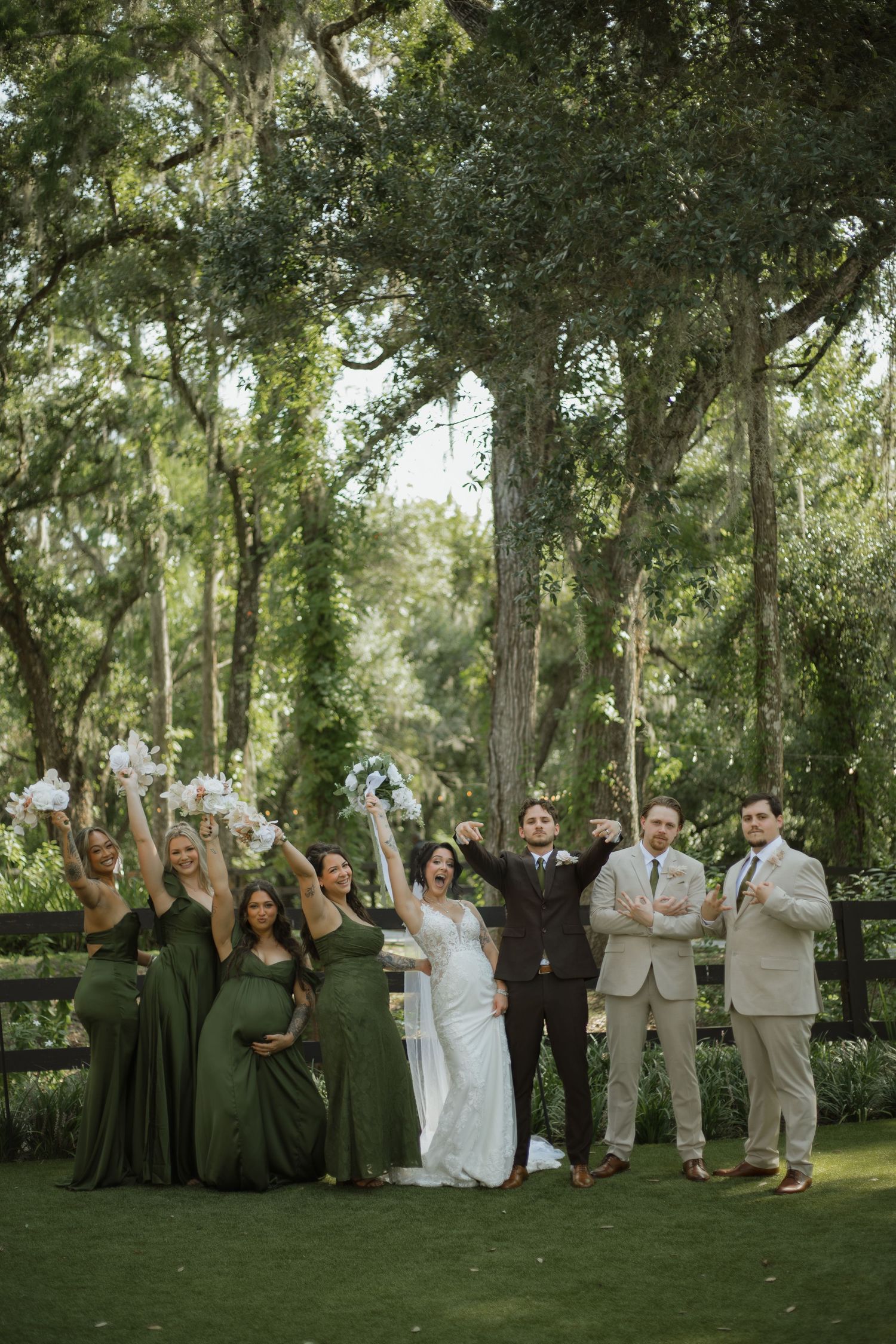 Bride and groom celebrate with bridesmaids in green dresses and groomsmen in tan suits under oak trees at an outdoor garden wedding.