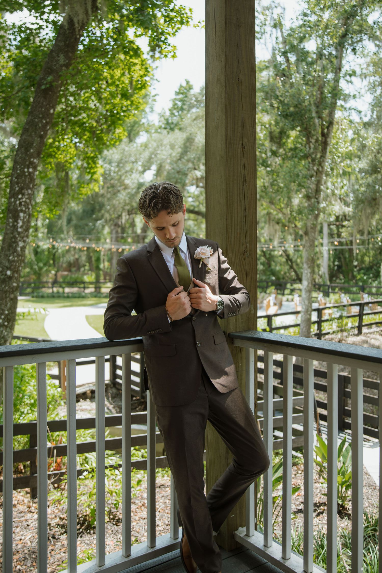 Groom in a brown suit with a floral boutonniere adjusts his jacket while leaning on a porch railing surrounded by lush green trees.