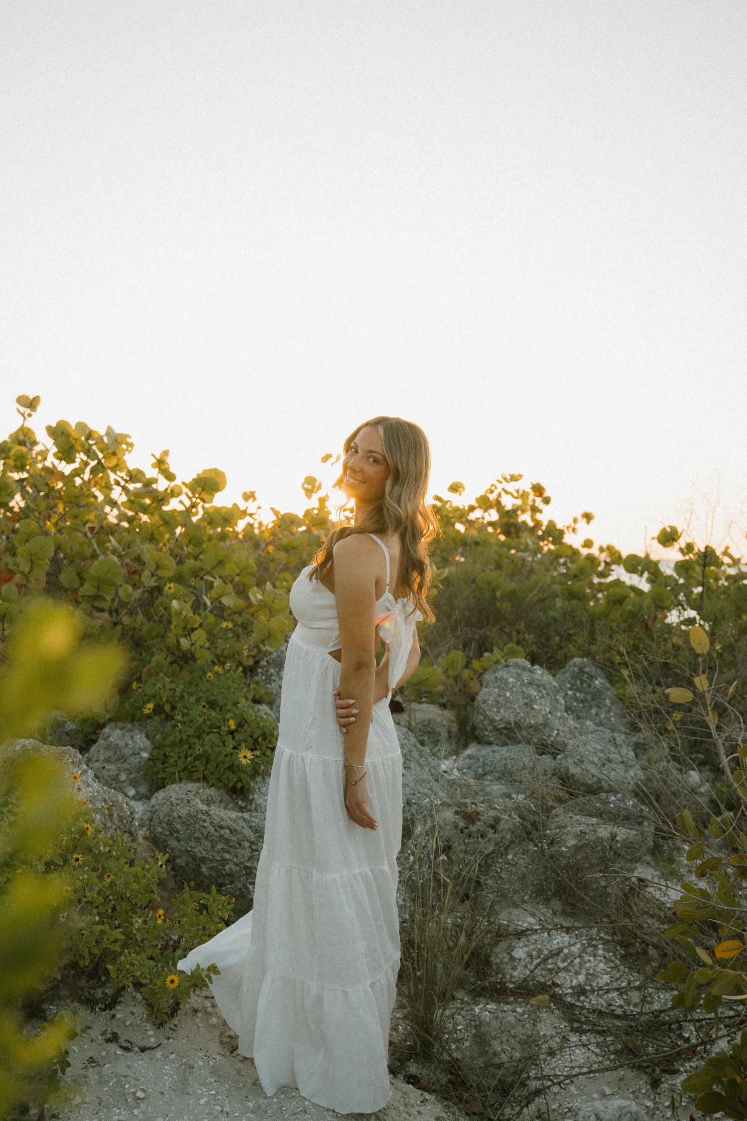 Woman in a long white tiered dress stands among rocks and coastal greenery as golden sunset light glows behind her.