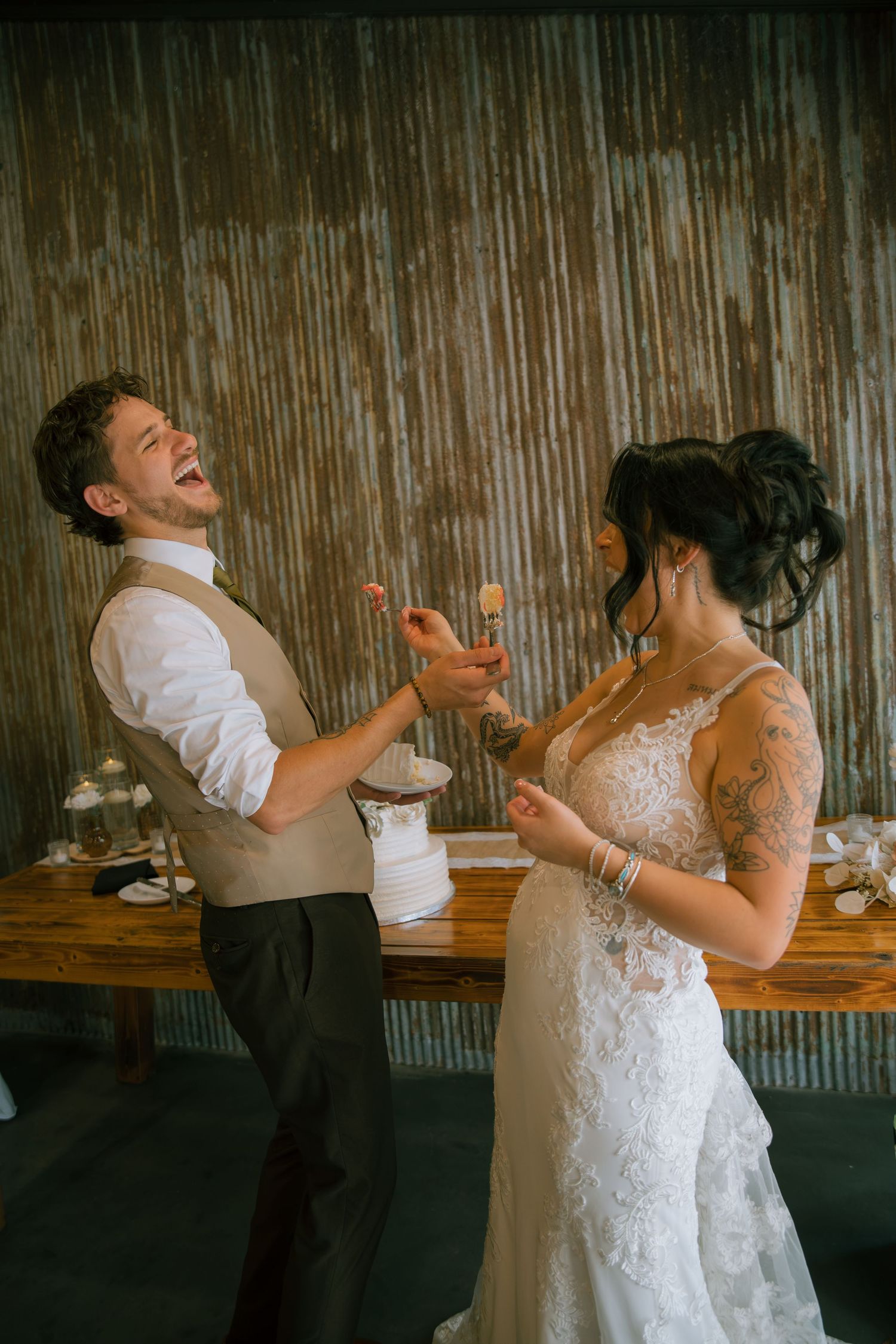 Groom in a tan vest laughs as the tattooed bride in a lace wedding gown playfully feeds him wedding cake.