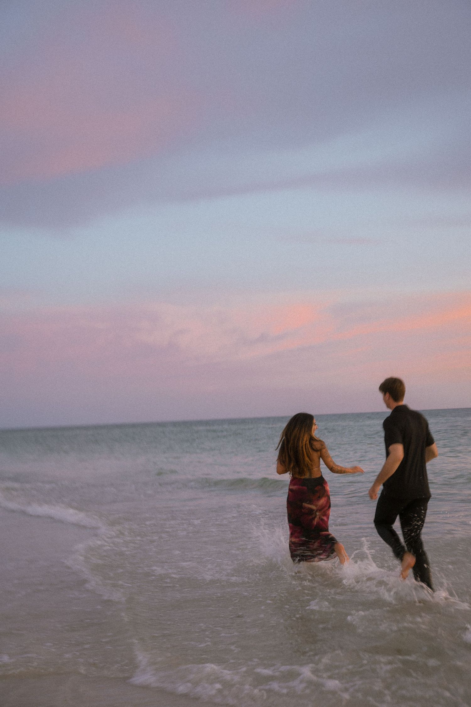 Couple holds hands and runs into shallow ocean waves at dusk beneath a soft pink and purple sky.