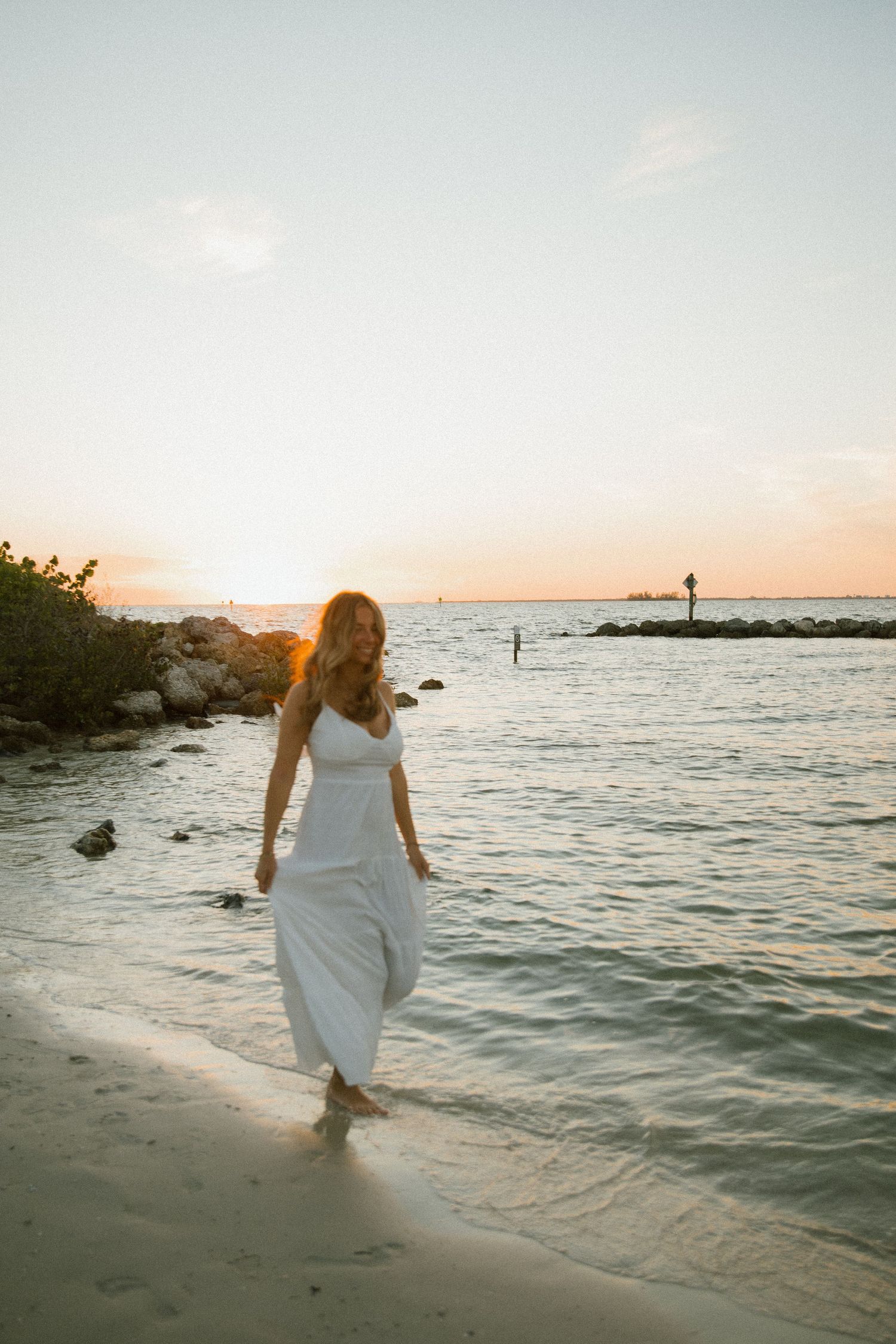 Woman in a white dress walks barefoot along the sandy shoreline as the sun sets over calm ocean water.