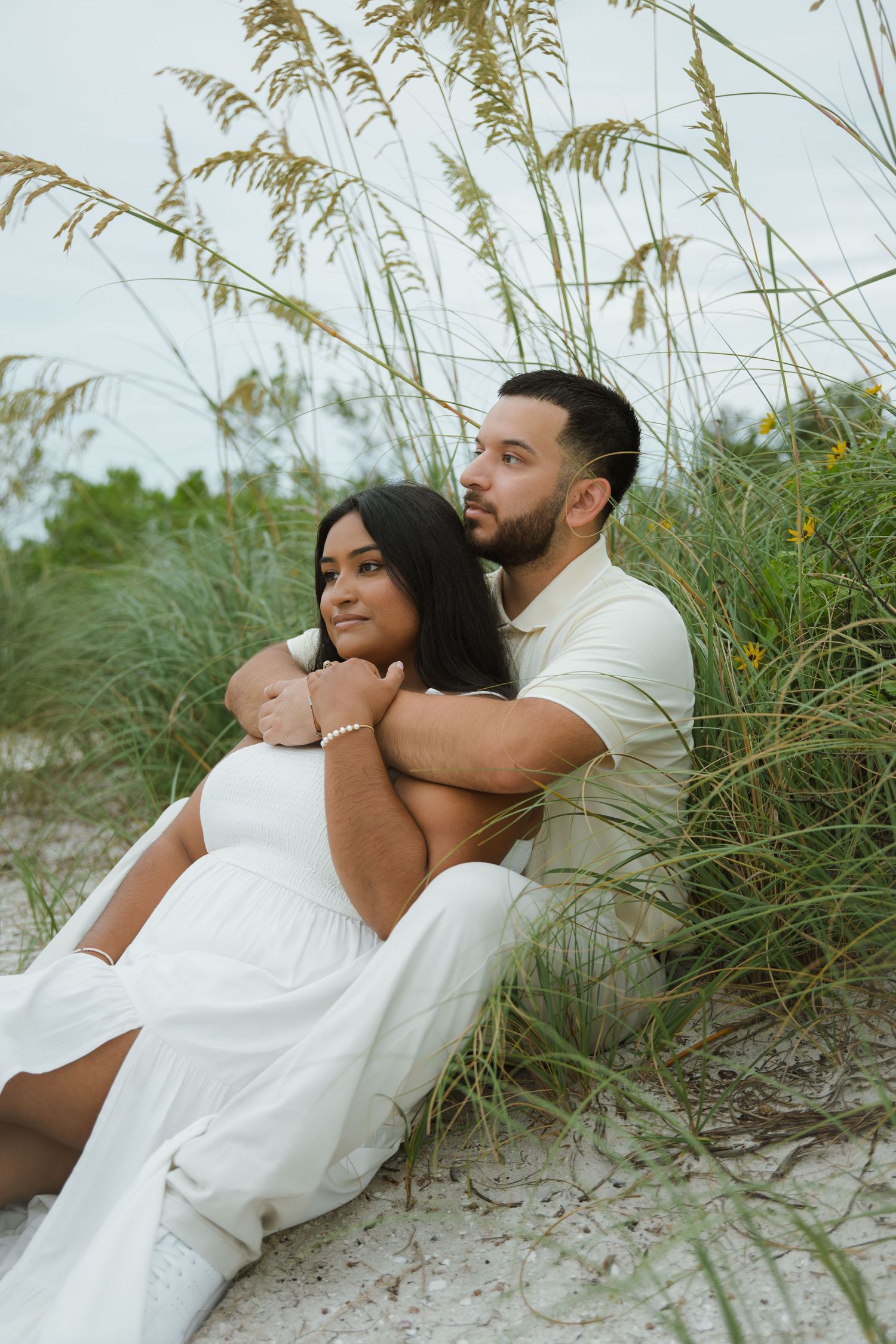Couple in white outfits sit close together on a sandy beach, embracing among tall sea oats during an outdoor engagement photo session.