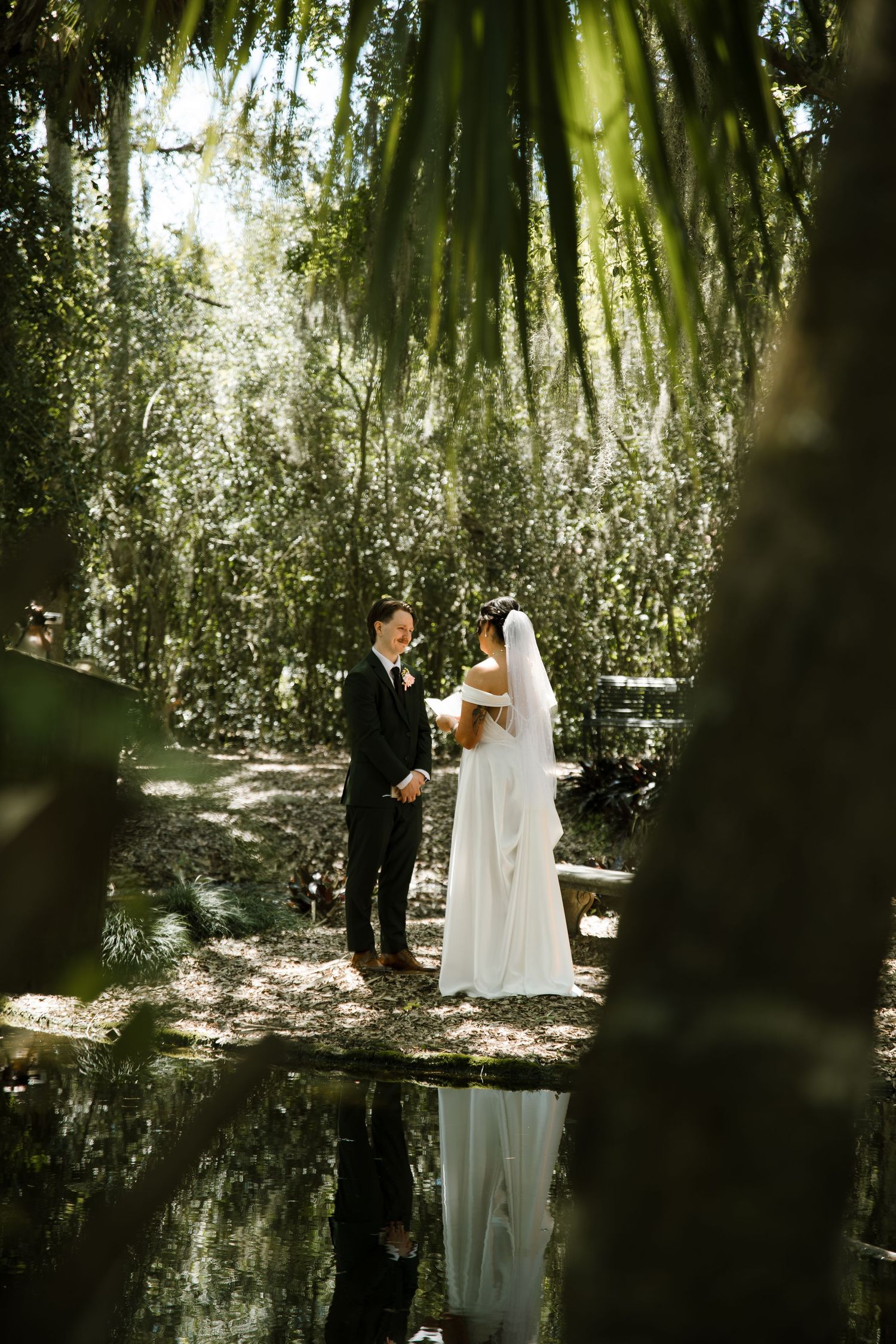 Bride and groom exchange vows in a sunlit wooded garden, their reflection shimmering in a still pond.
