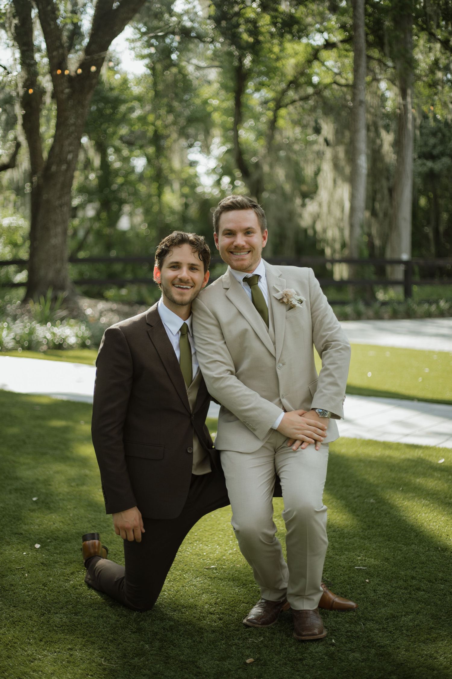 Groom in a brown suit kneels beside a smiling groomsman in a tan suit with a boutonniere on a green lawn at an outdoor wedding.