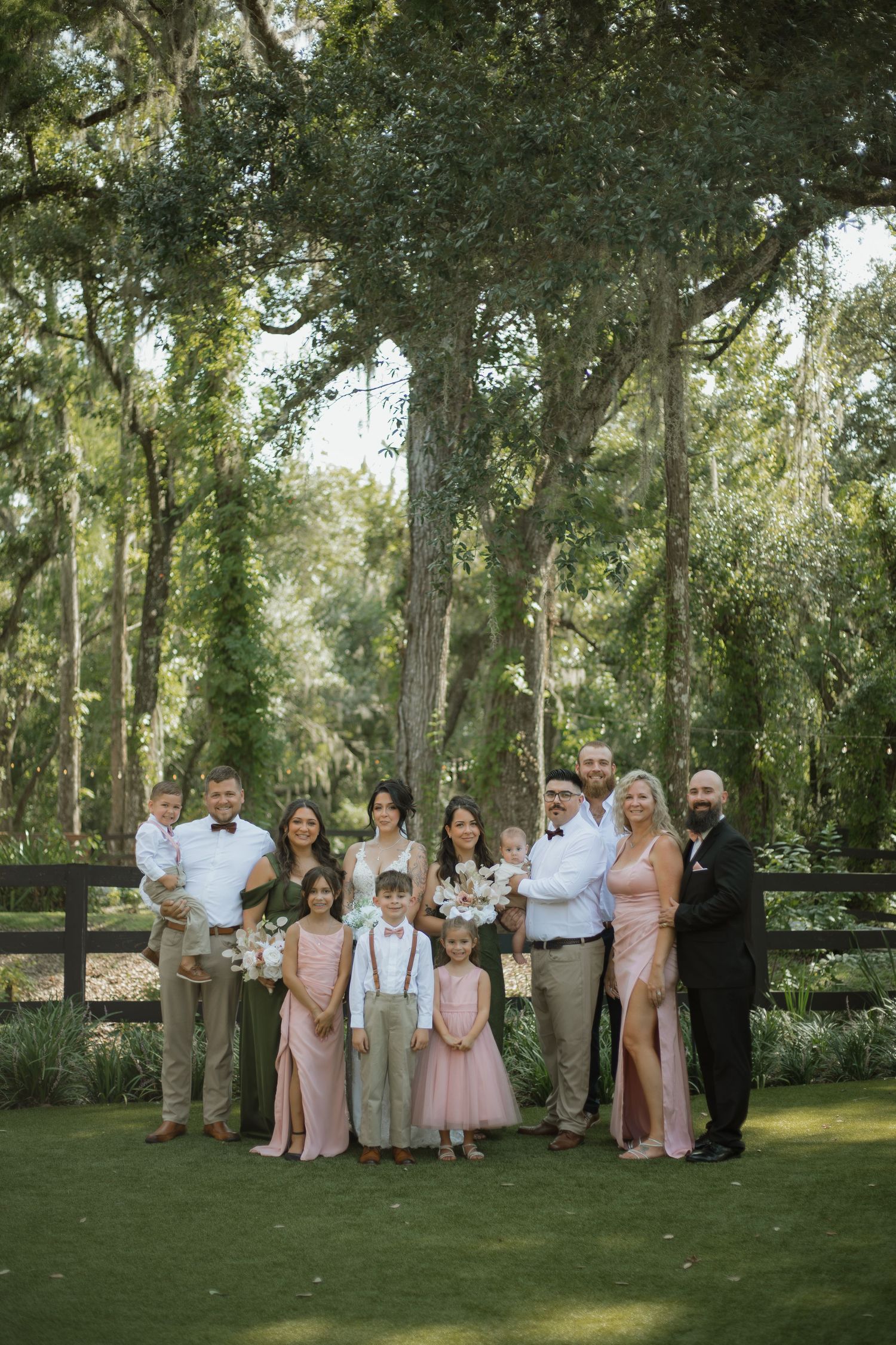 Wedding family portrait with the bride, groom, flower girls, children and relatives posed together on a lawn beneath tall trees.